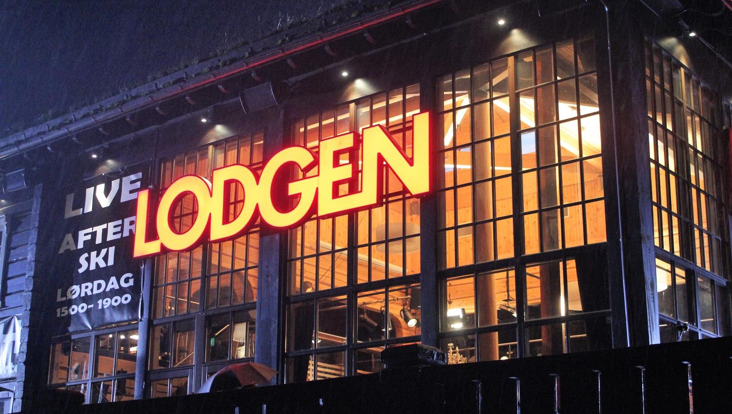 Kveldsbilde av bygning med store vinduer og rødt, opplyst skilt «LODGEN», med varmt lys inne..
