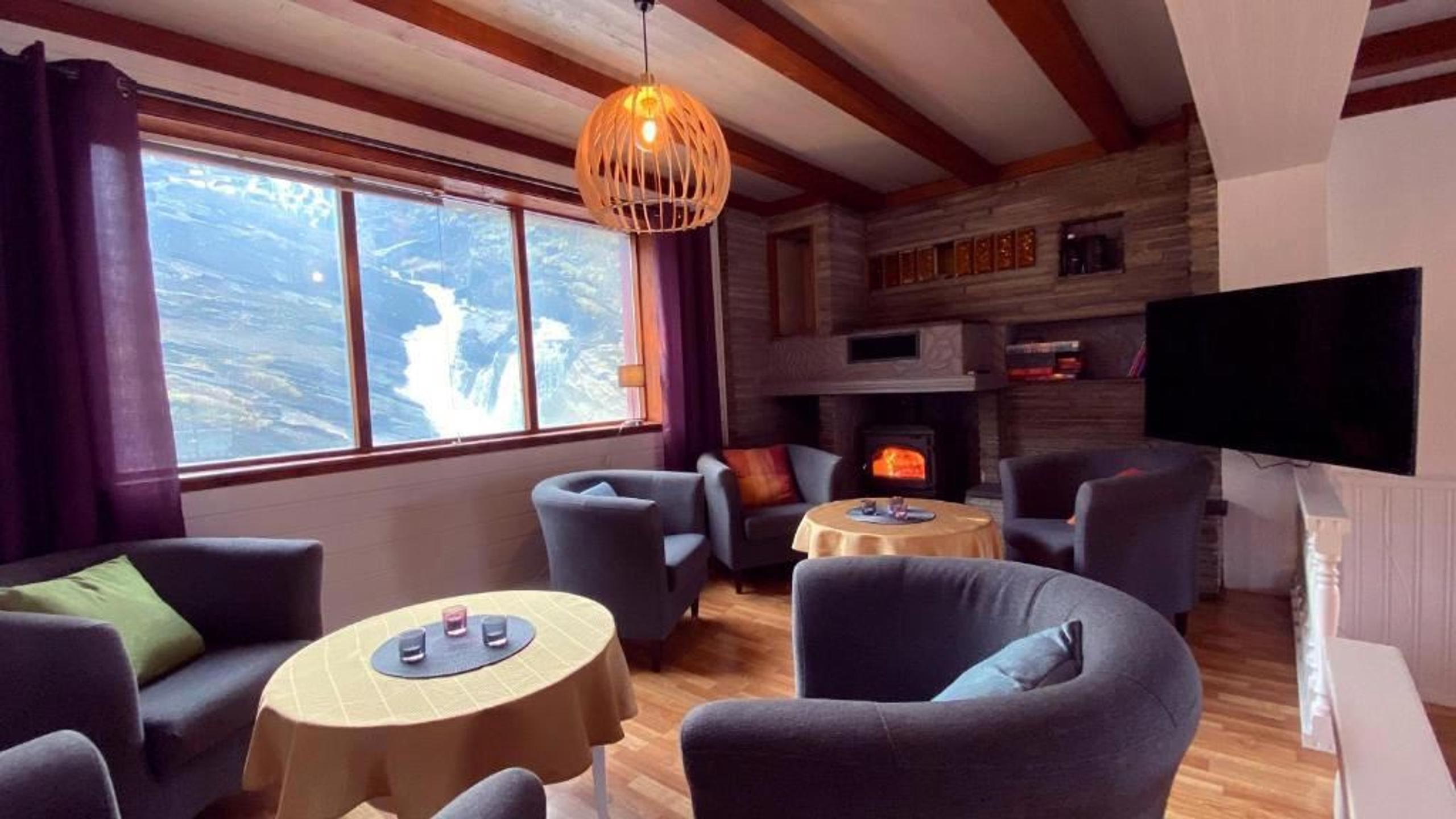 Warm living room with armchairs, round tables, a glowing stove, and large window showing snowy mountains outside.