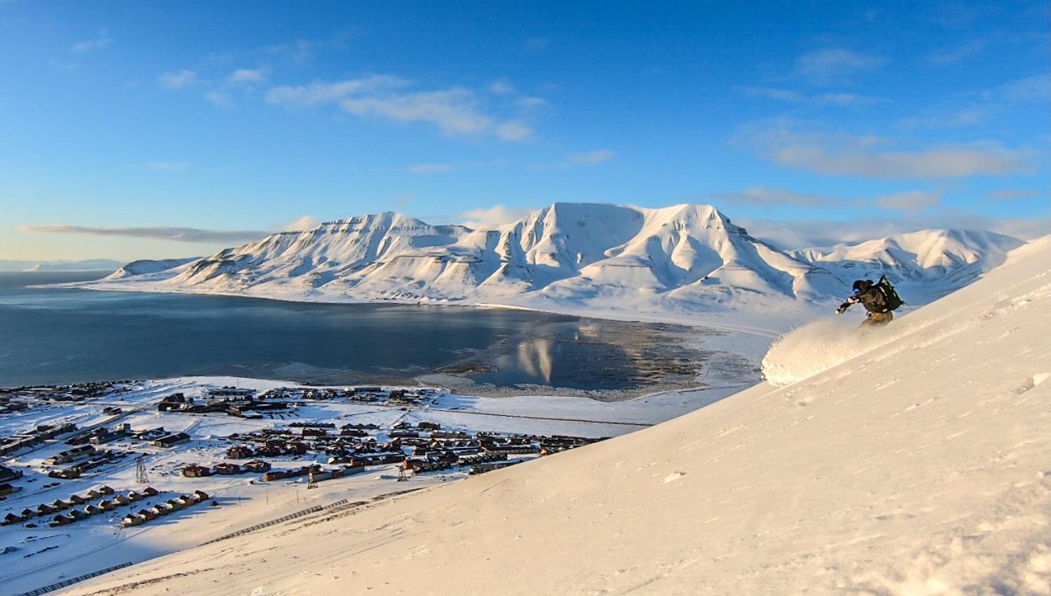 A person skiing down a mountain with Longyearbyen and a mountainous landscape in the background