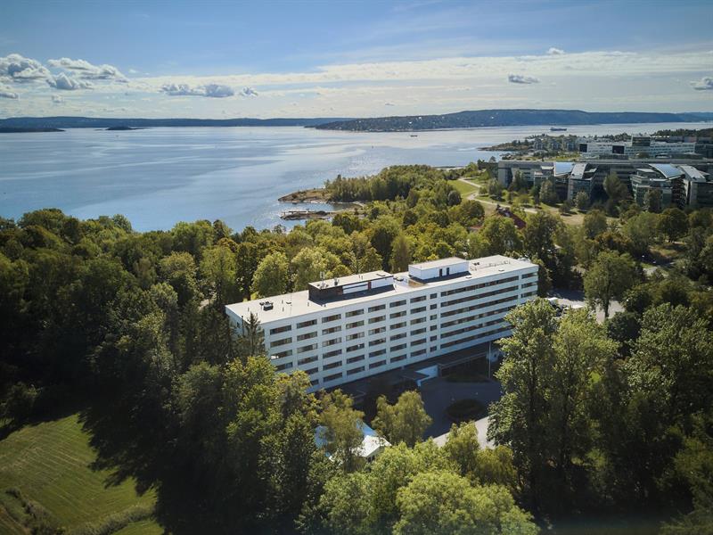 Luftfoto av hotellbygning omgitt av grøntområde ved sjøen med utsikt over fjorden.