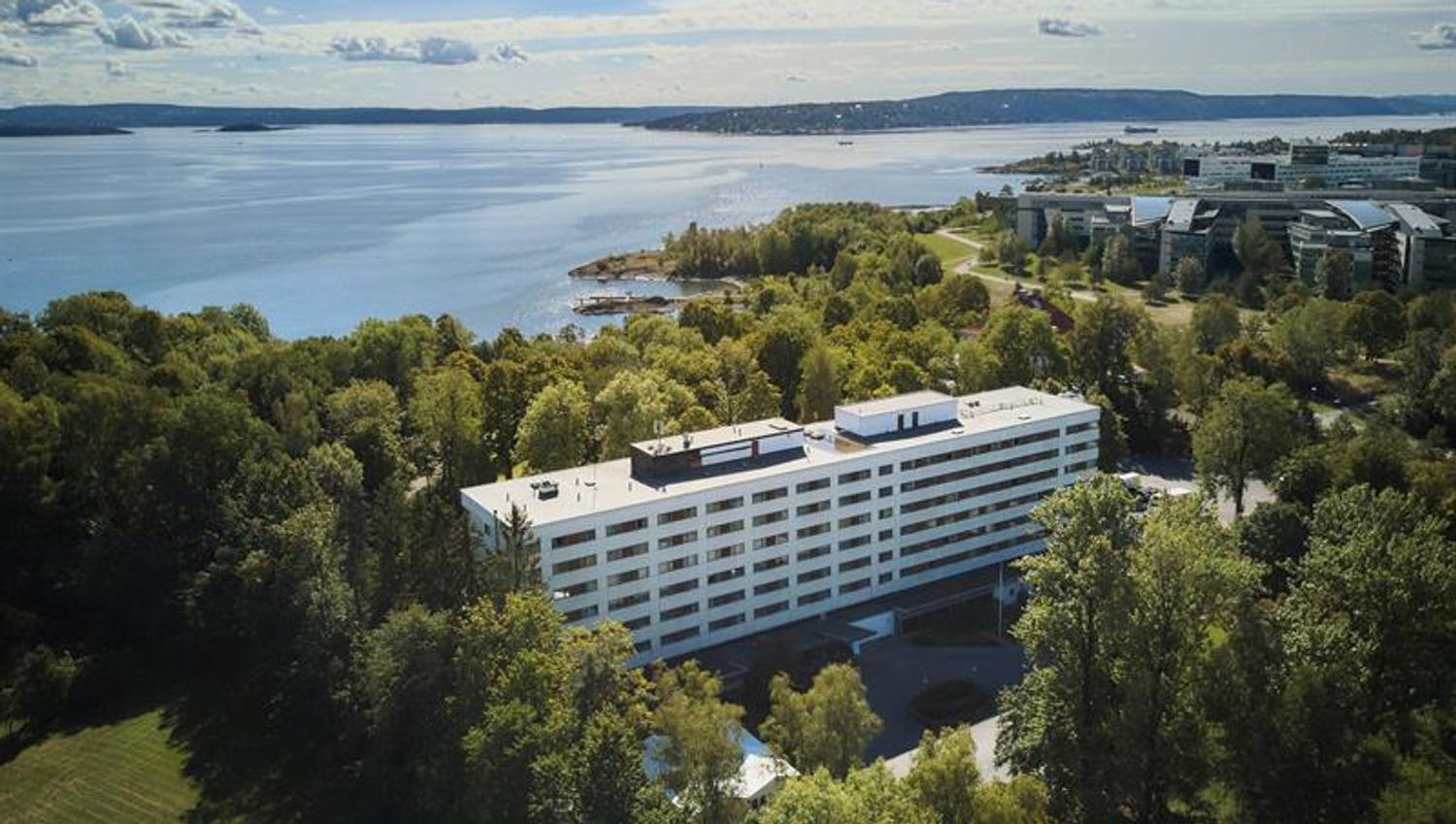 Luftfoto av hotellbygning omgitt av grøntområde ved sjøen med utsikt over fjorden.