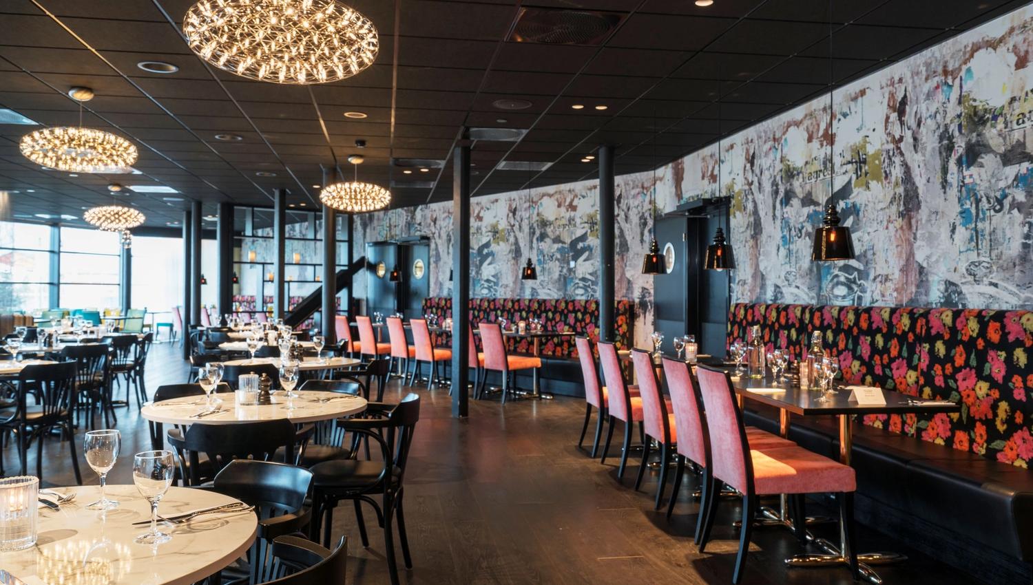 En moderne restaurant med runde og avlange bord, dekorative lamper og fargerike sitteplasser.
