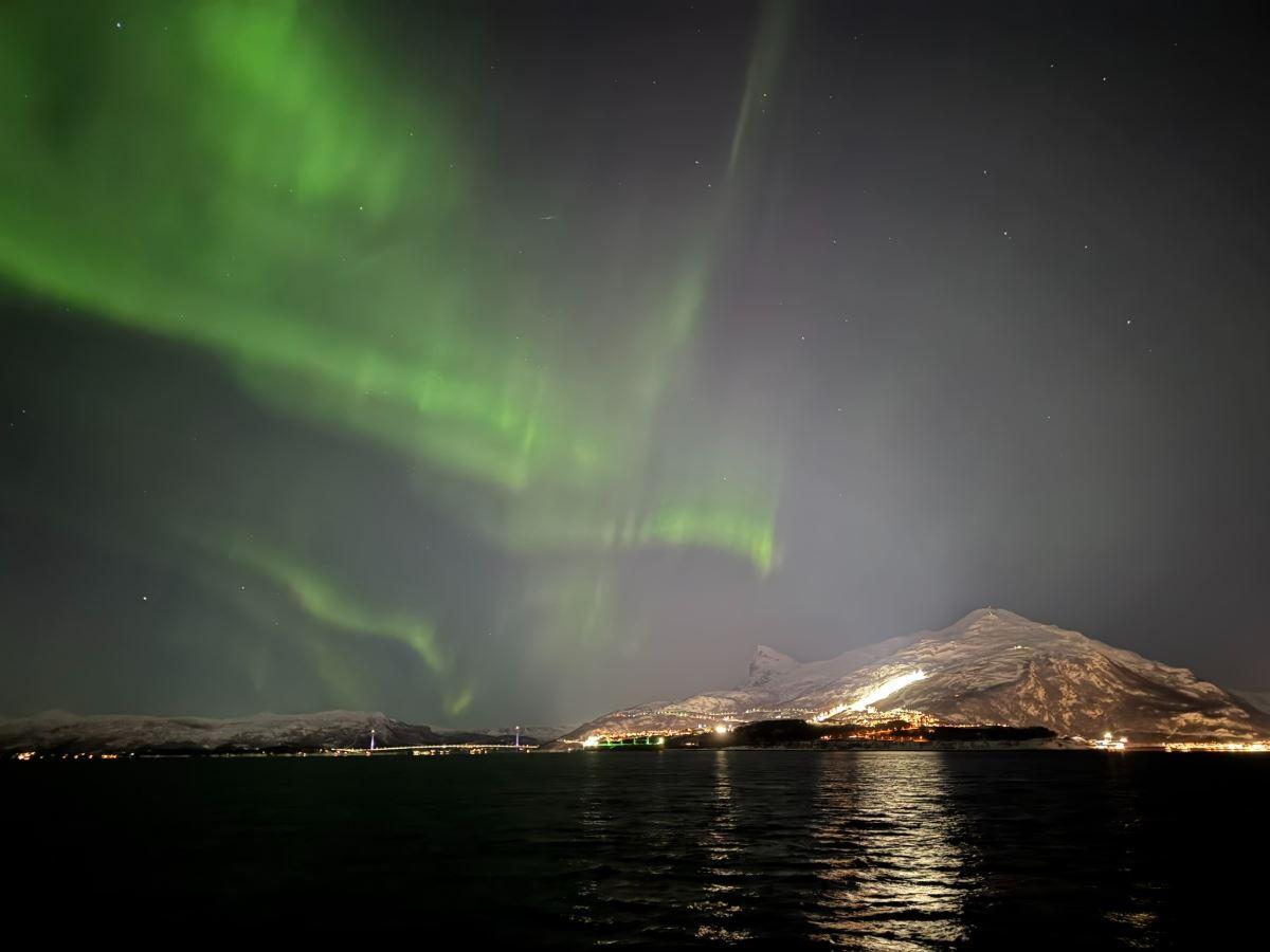 Green aurora curtains glow above a starry night sky, with a snow-covered mountain and lights reflecting on dark water.