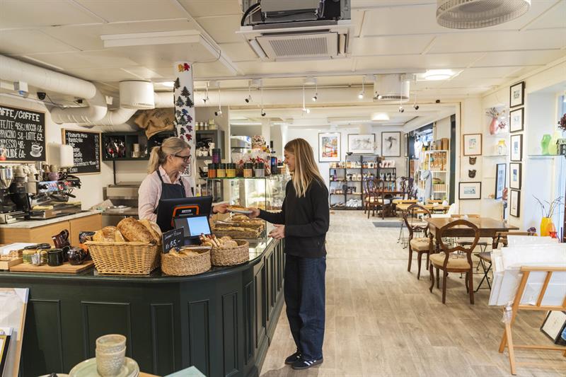 A woman buys freshly baked goods at the counter of Iversen Bakeri & Galleri in Norheimsund, surrounded by baskets of bread, a coffee machine and artwo
