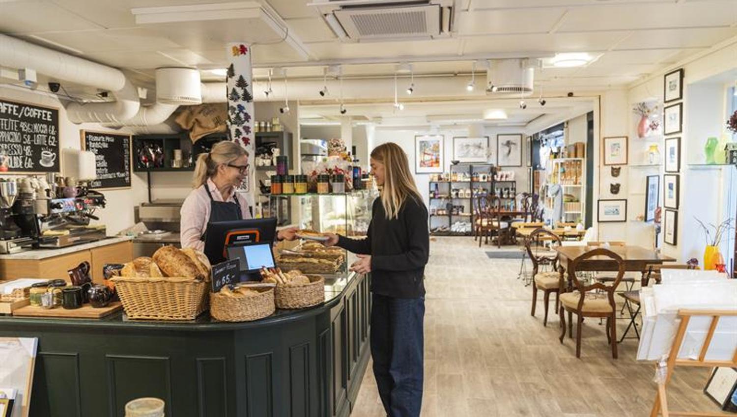 A woman buys freshly baked goods at the counter of Iversen Bakeri & Galleri in Norheimsund, surrounded by baskets of bread, a coffee machine and artwo