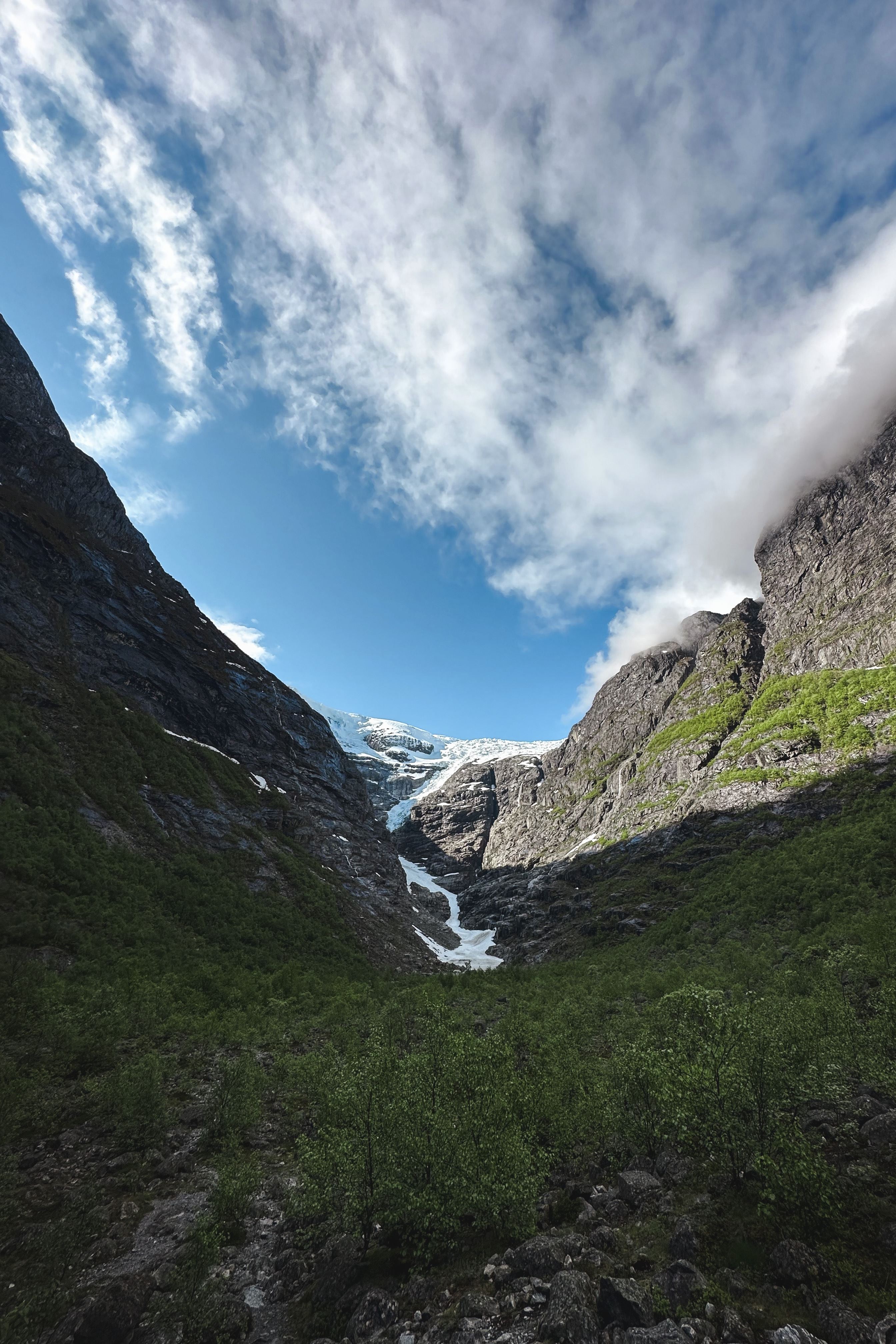 Rocky mountain valley with green shrubs, steep cliffs, and a distant glacier beneath a bright blue sky with clouds..