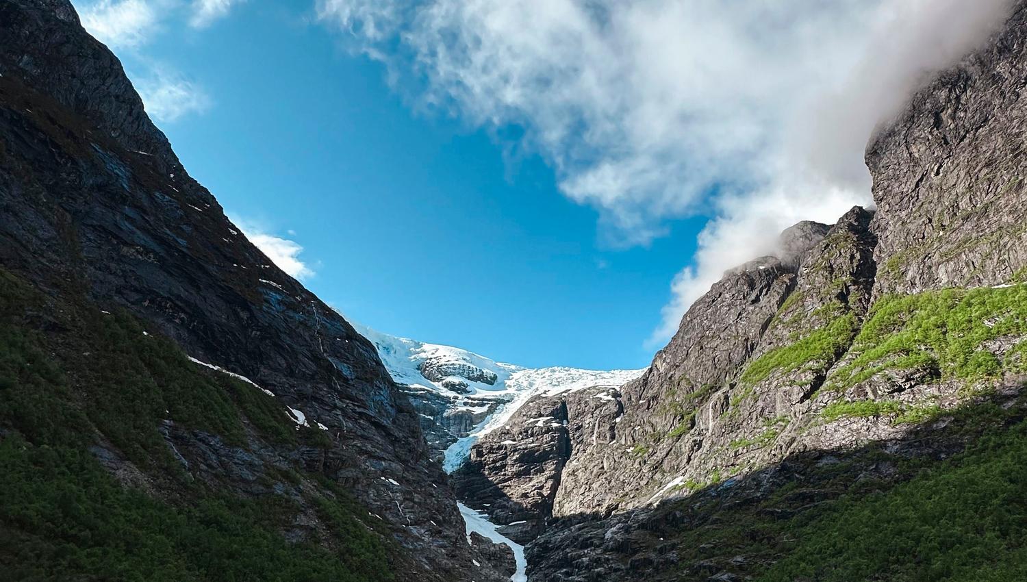 Rocky mountain valley with green shrubs, steep cliffs, and a distant glacier beneath a bright blue sky with clouds..
