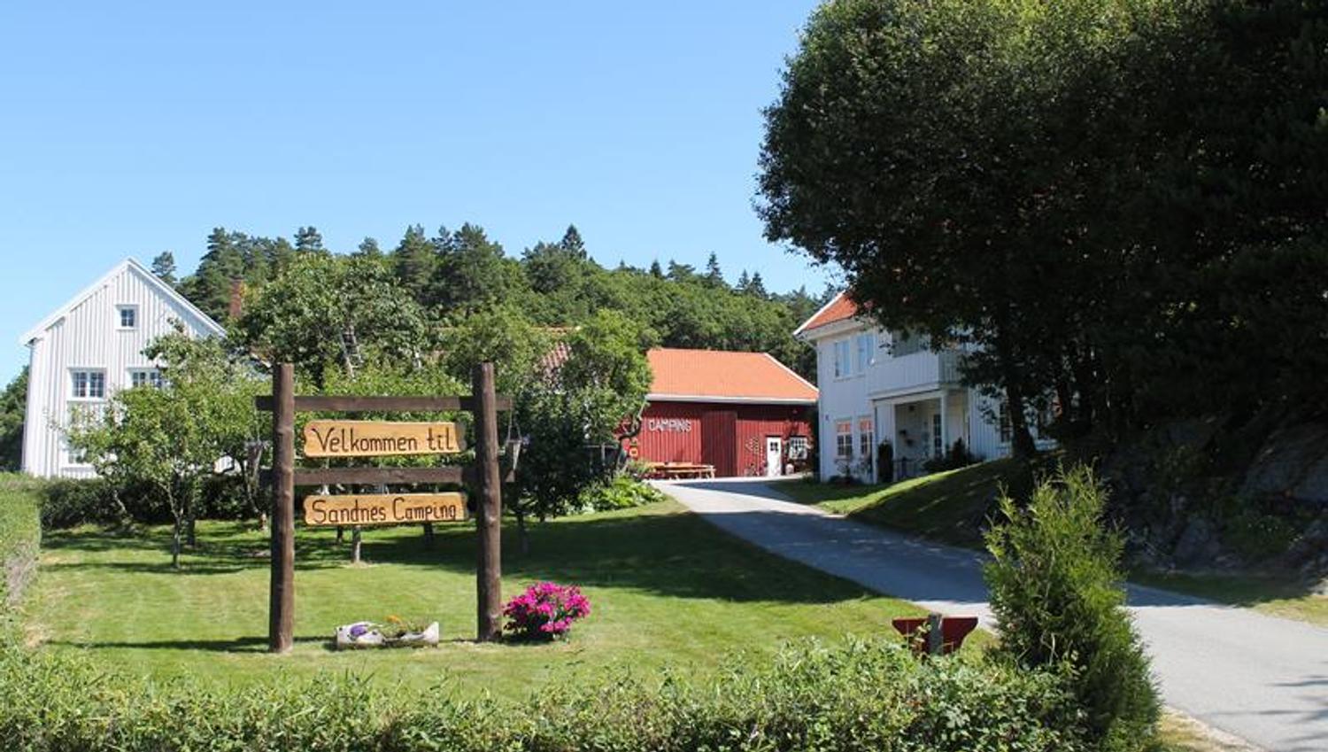 Einfahrt zum Sandnes Camping. Ein großes Holzschild mit der Aufschrift „Willkommen bei Sandnes Camping“ steht in einem Garten.
