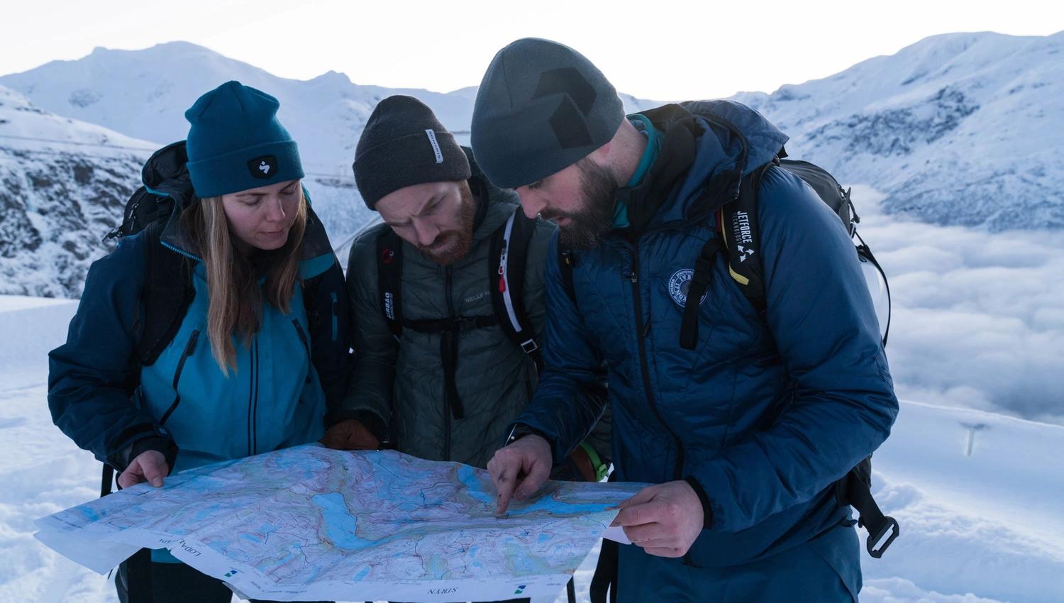 Three bundled hikers with backpacks study a large paper map on a snowy ridge, with mountains and clouds behind.
