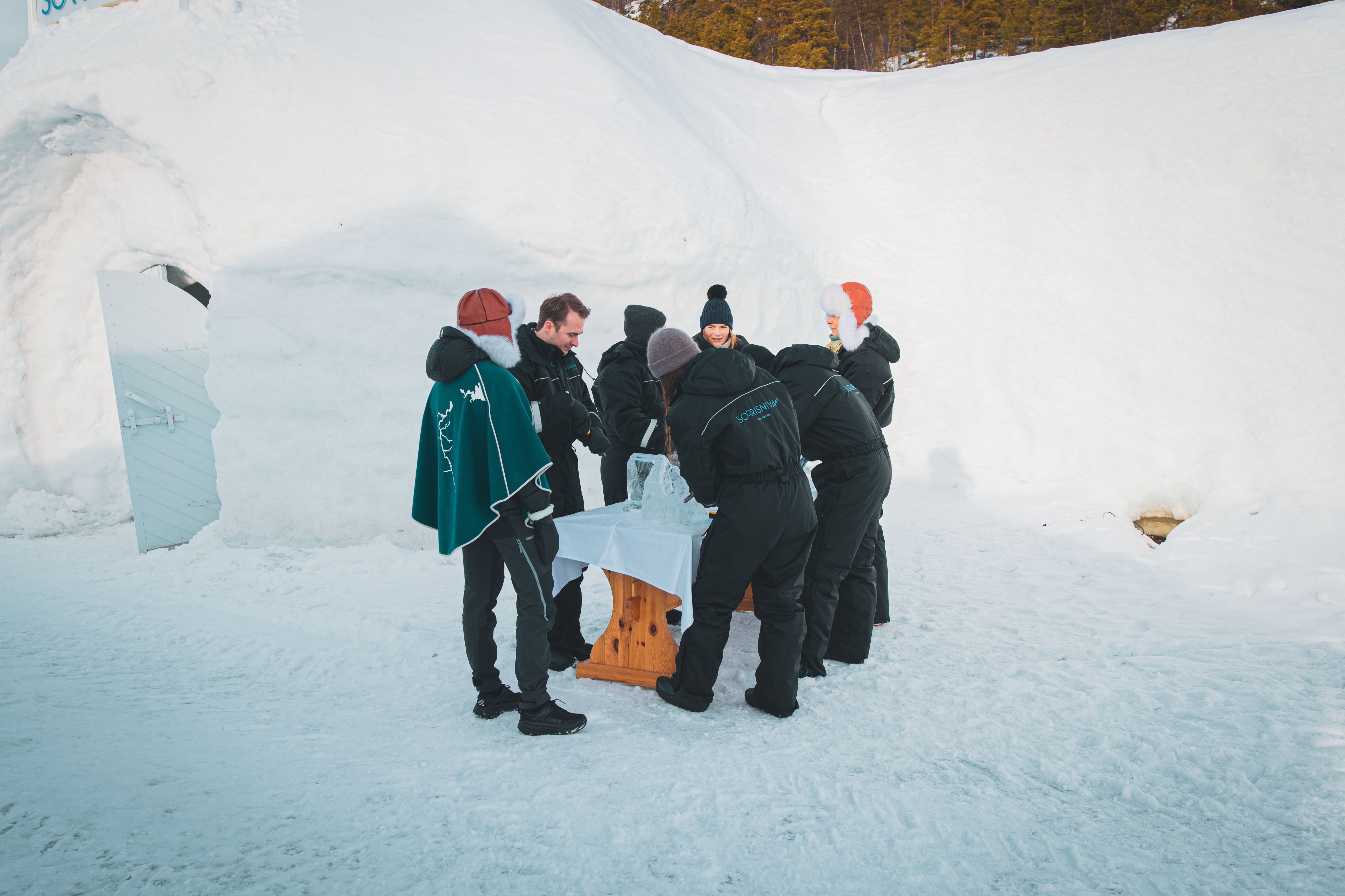 Group of people in winter clothes gathered around a table in a snowy landscape.
