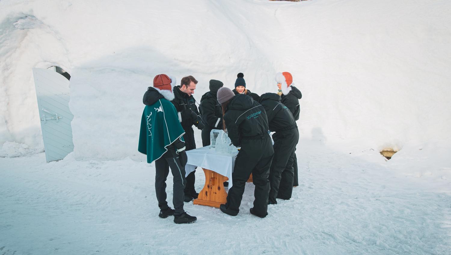 Group of people in winter clothes gathered around a table in a snowy landscape.
