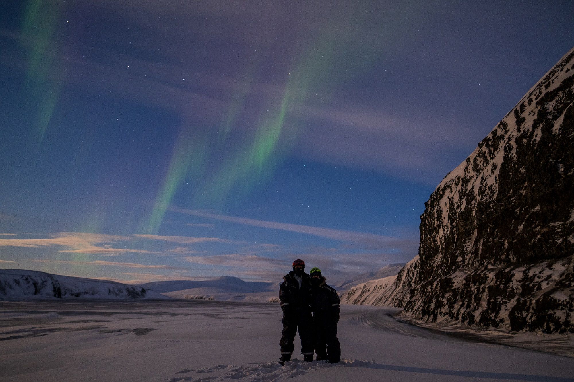 Two persons in snowmobile equipment hugging with northern lights in the skies above