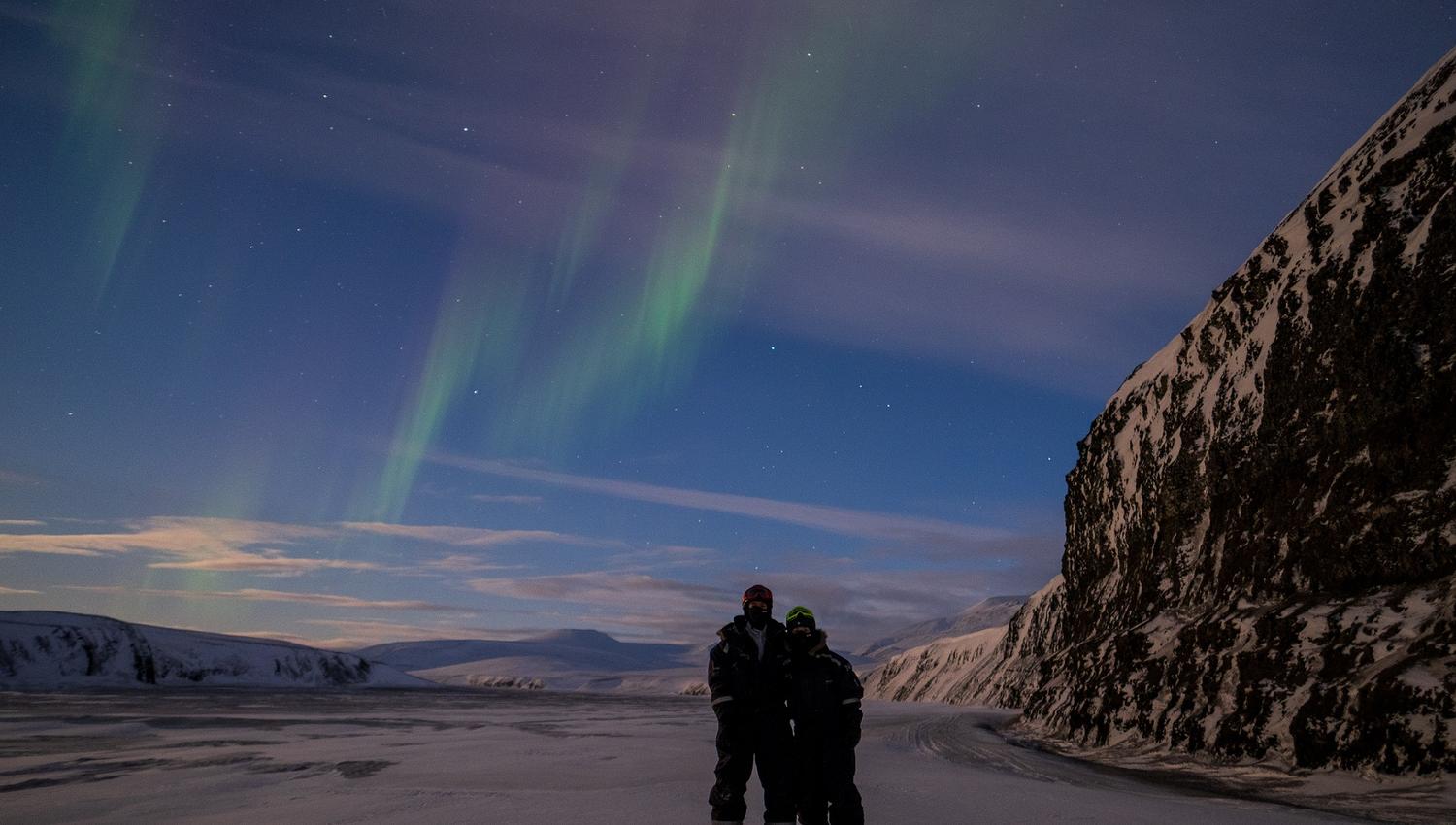 Two persons in snowmobile equipment hugging with northern lights in the skies above