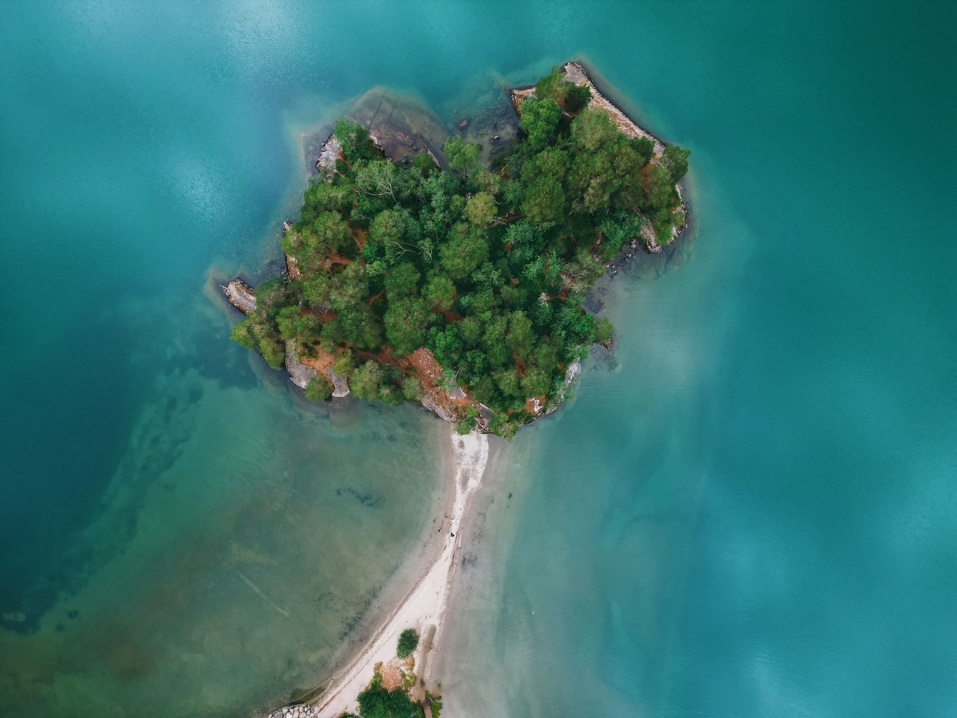 Aerial view of a small tree-covered island surrounded by turquoise water, connected by a narrow sandy sandbar..