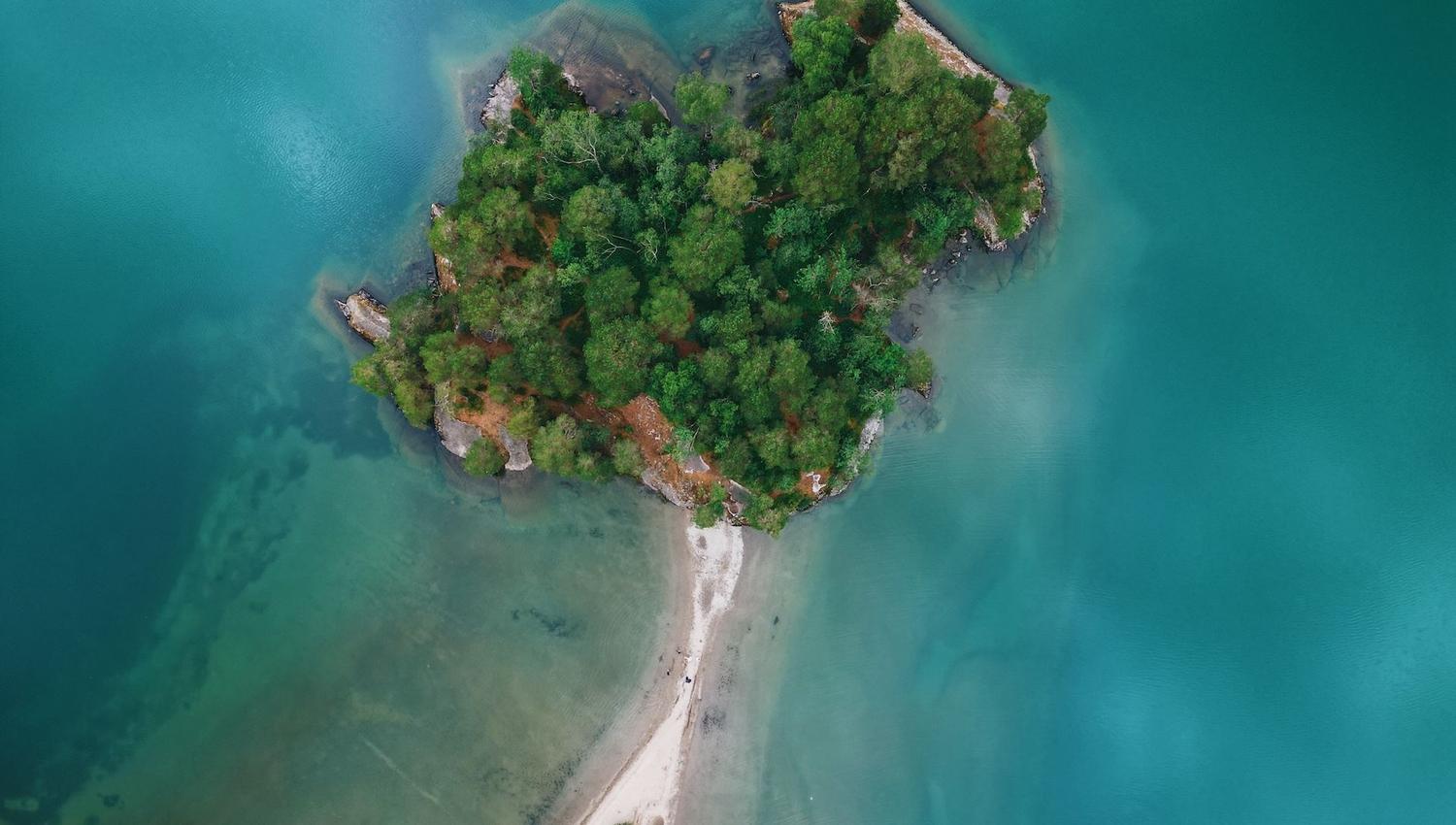 Aerial view of a small tree-covered island surrounded by turquoise water, connected by a narrow sandy sandbar..