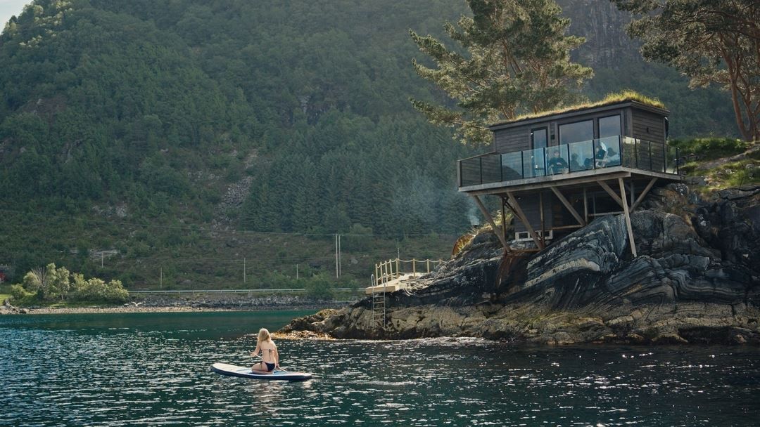 A person kneels on a paddleboard in calm water near a modern cabin on rocky cliffs with forested hills behind..