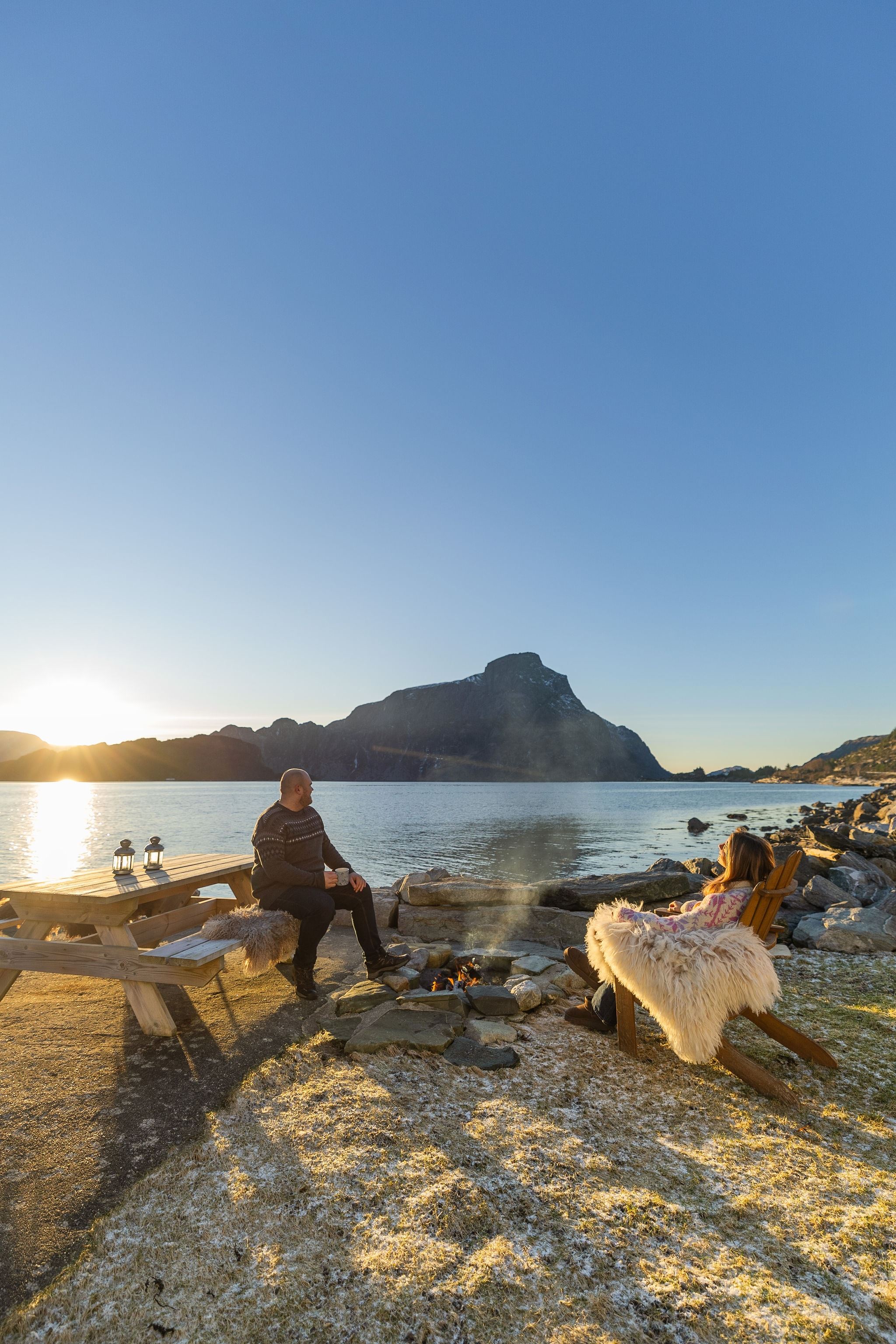 Two people relax by a small campfire near a calm lake, with a picnic table, rocks, and distant mountains at sunset at the Seahouse at Hornelen Panorama in Bremanger..