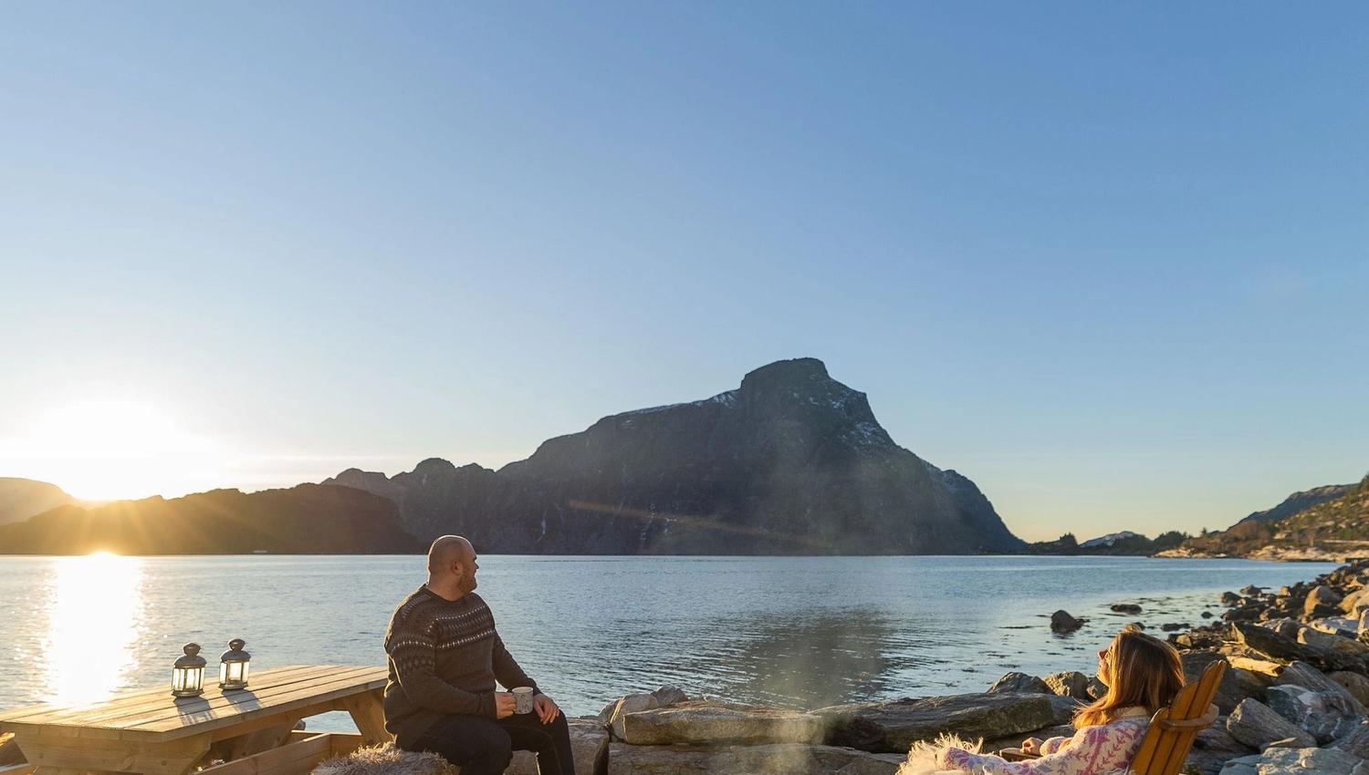 Two people relax by a small campfire near a calm lake, with a picnic table, rocks, and distant mountains at sunset at the Seahouse at Hornelen Panorama in Bremanger..
