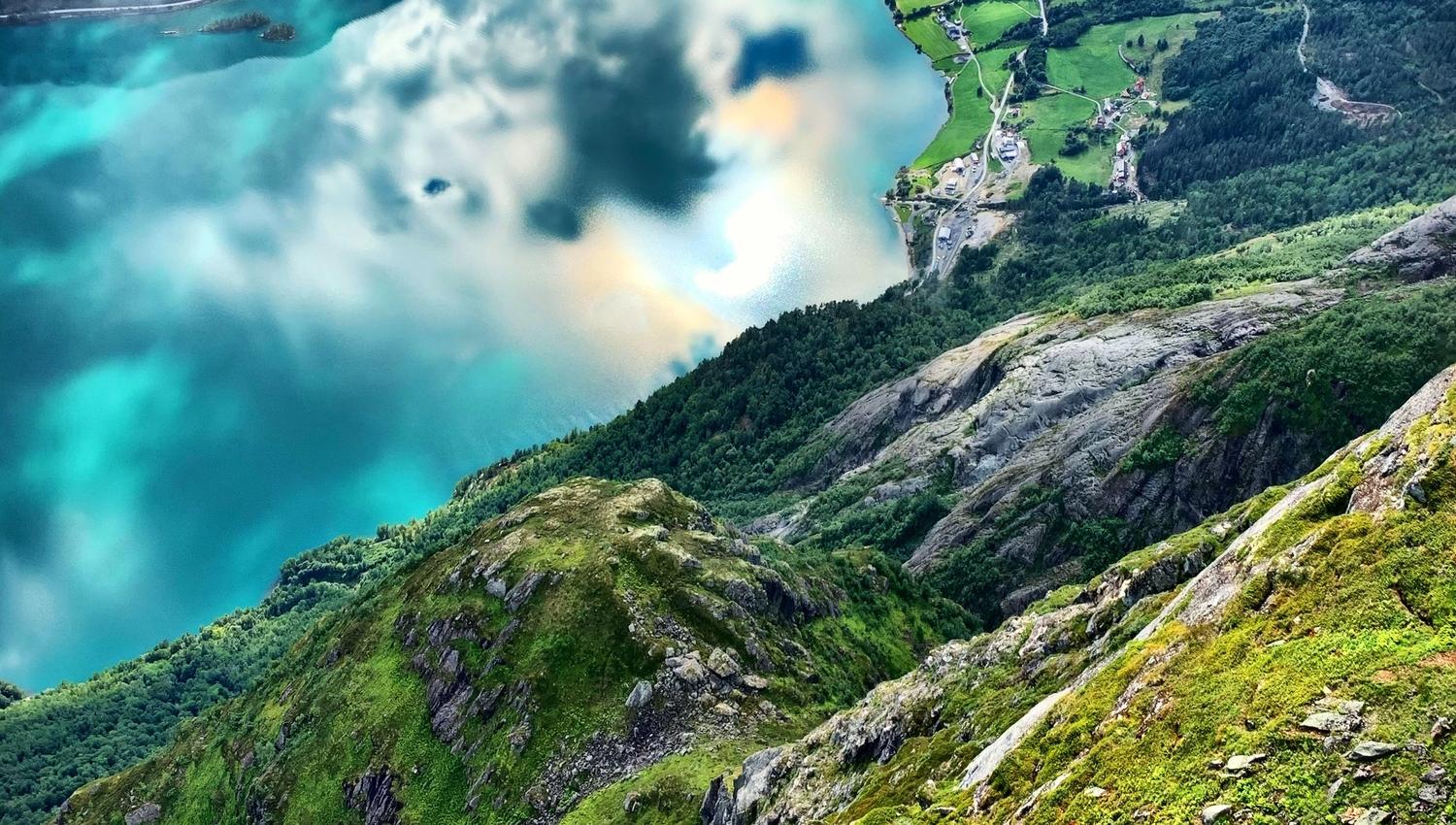 High view from a grassy rocky mountainside over a turquoise lake, with clouds, forested slopes, and small farms..