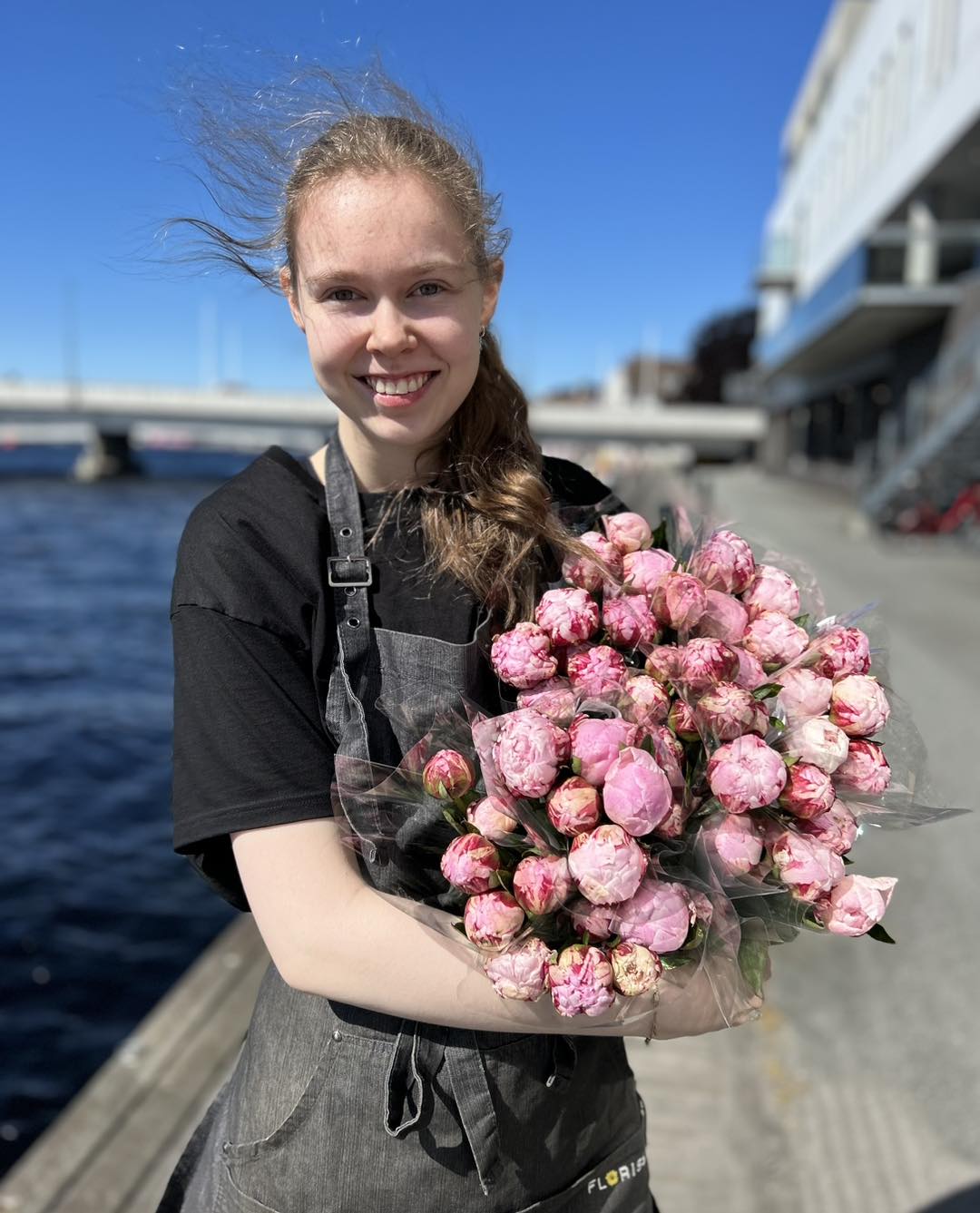 Smilende ansatt fra blomsterbutikken Floriss, viser frem flotte blomster, med Mandals elva i bakgrunnen