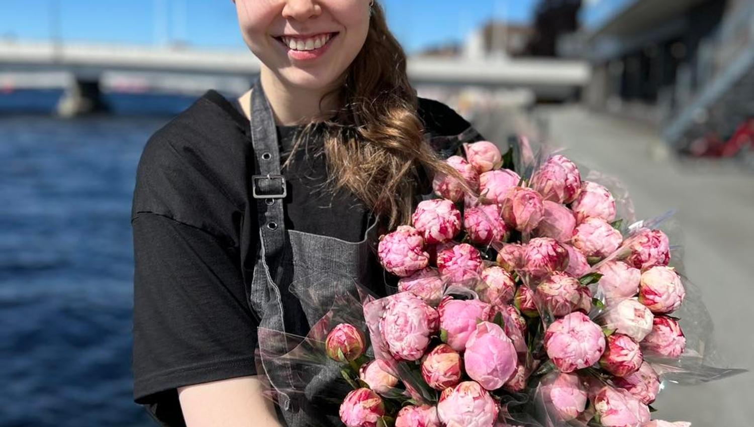 Smilende ansatt fra blomsterbutikken Floriss, viser frem flotte blomster, med Mandals elva i bakgrunnen