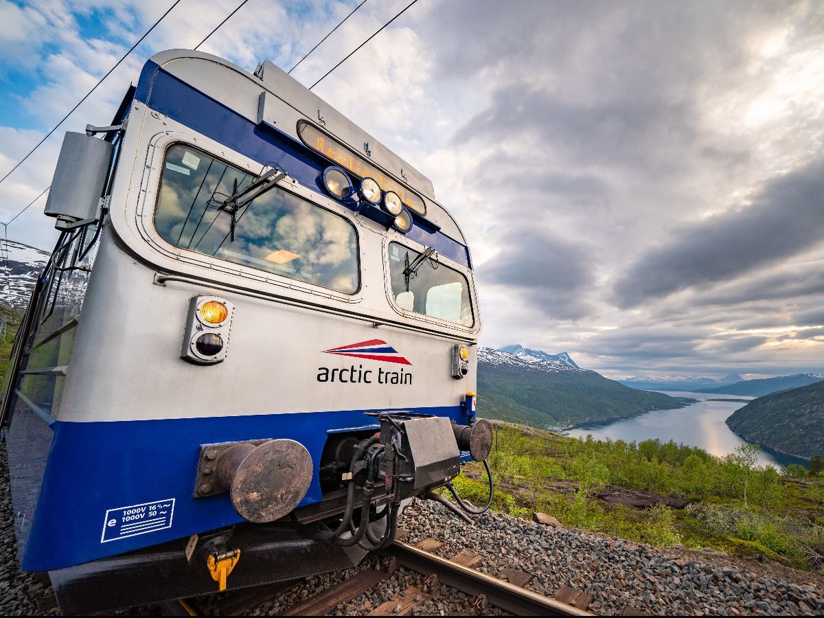Nærbilde av fronten på et blåhvitt tog på skinnegang, med fjord, grønne åser og skyet himmel i bakgrunnen..