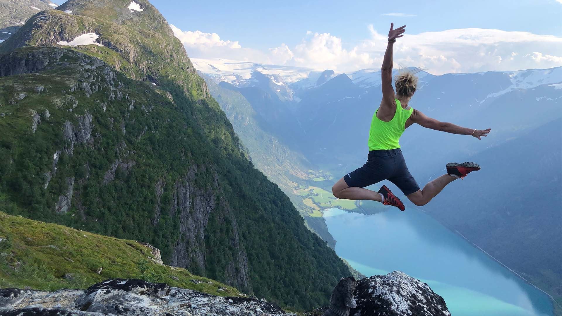 Person in a neon green top jumps near a rocky cliff with steep mountains and a blue fjord below..