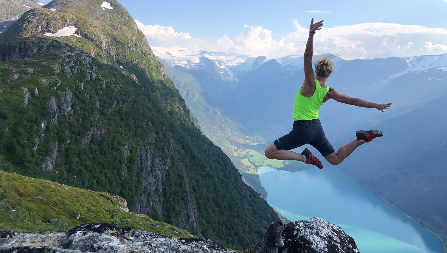 Person in a neon green top jumps near a rocky cliff with steep mountains and a blue fjord below..