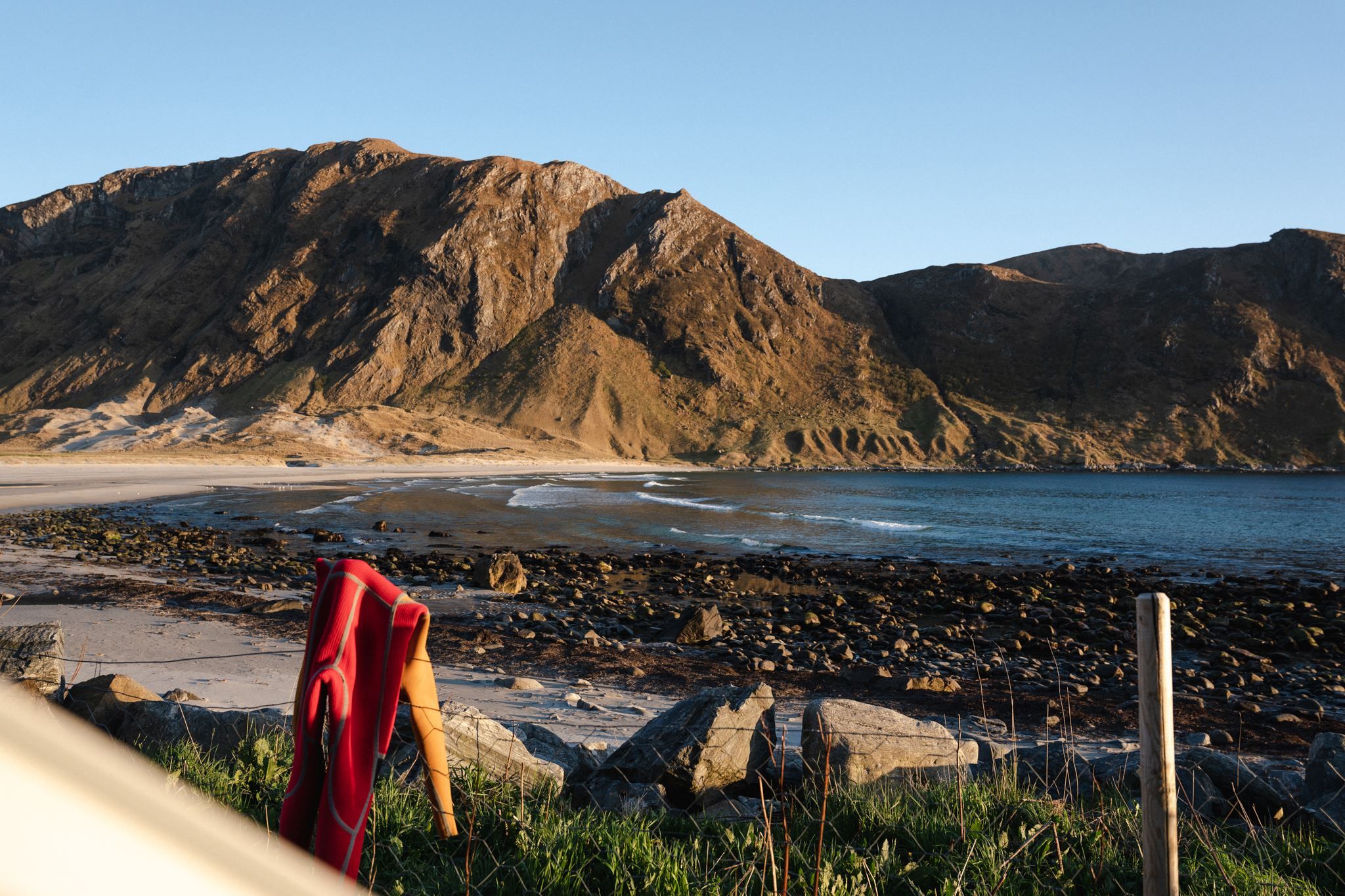 A red wetsuit draped over a post on grassy shore, facing a rocky beach, calm sea, and sunlit mountain ridge.