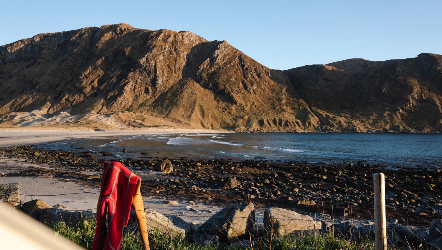 A red wetsuit draped over a post on grassy shore, facing a rocky beach, calm sea, and sunlit mountain ridge.