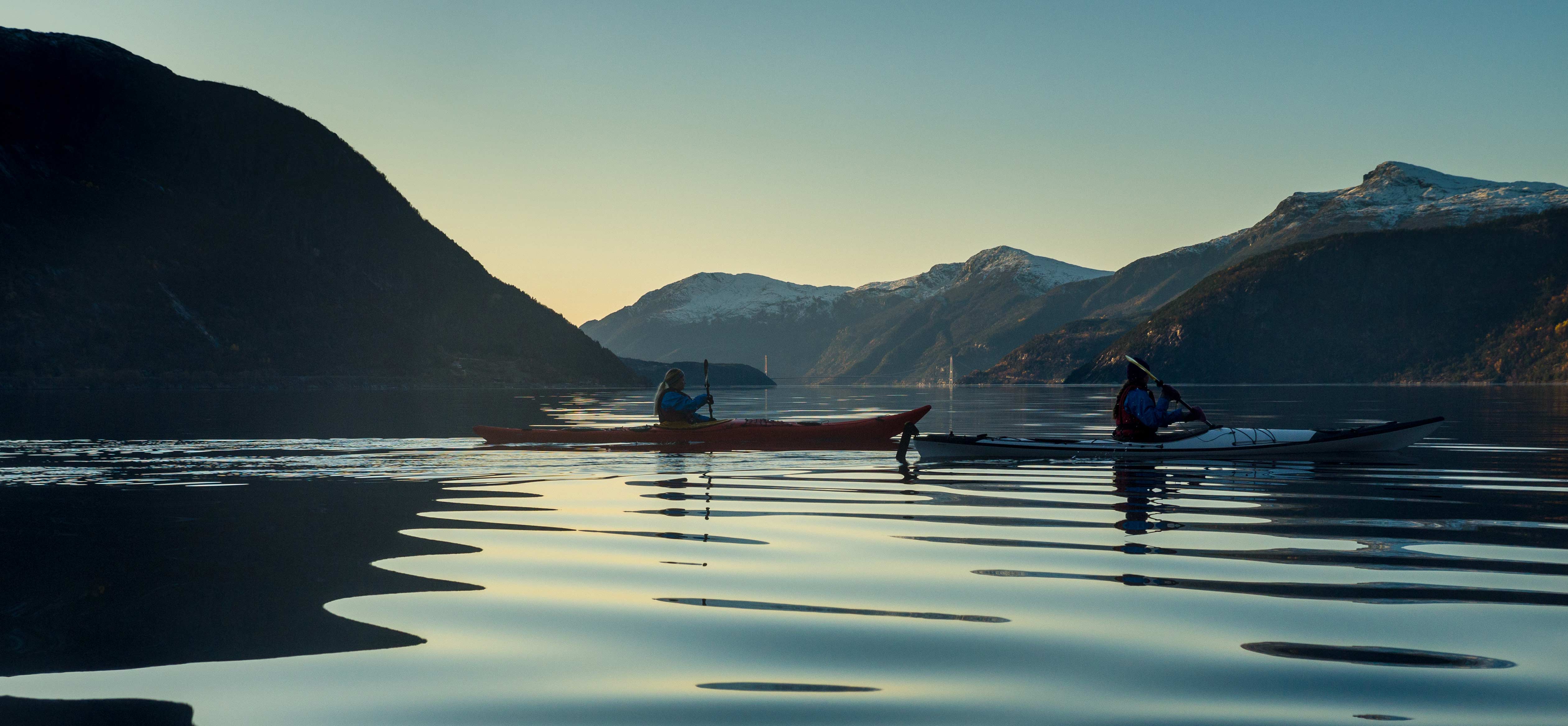 Kayakers on calm waters in the Hardangerfjord at sunset, with mountain silhouettes and reflections on the water.