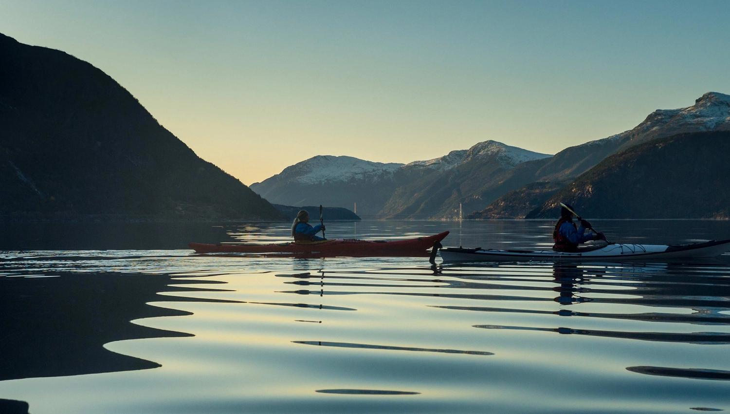 Kayakers on calm waters in the Hardangerfjord at sunset, with mountain silhouettes and reflections on the water.