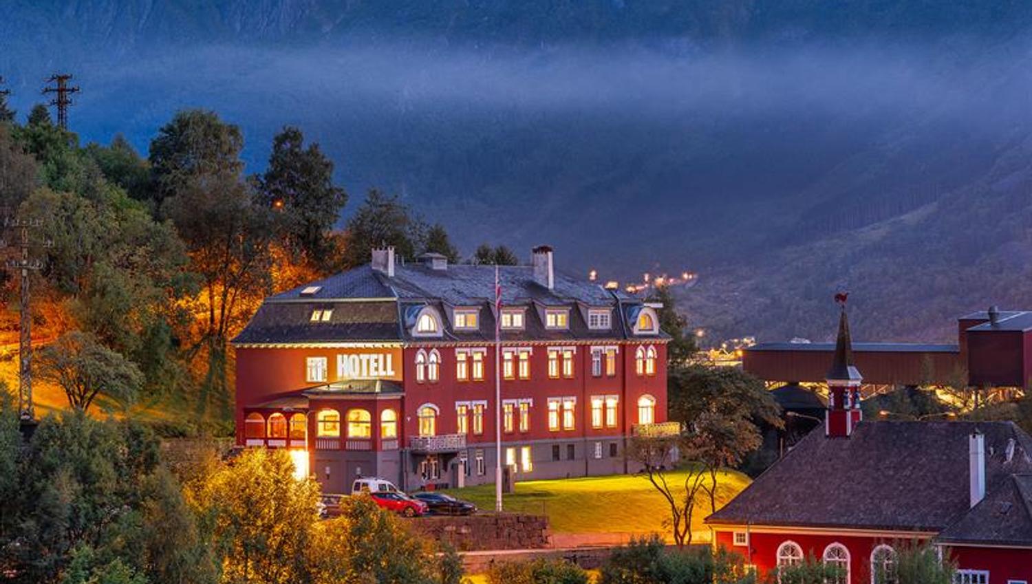 Historic Tyssedal Hotel illuminated at dusk, surrounded by mountains and the Hardangerfjord.