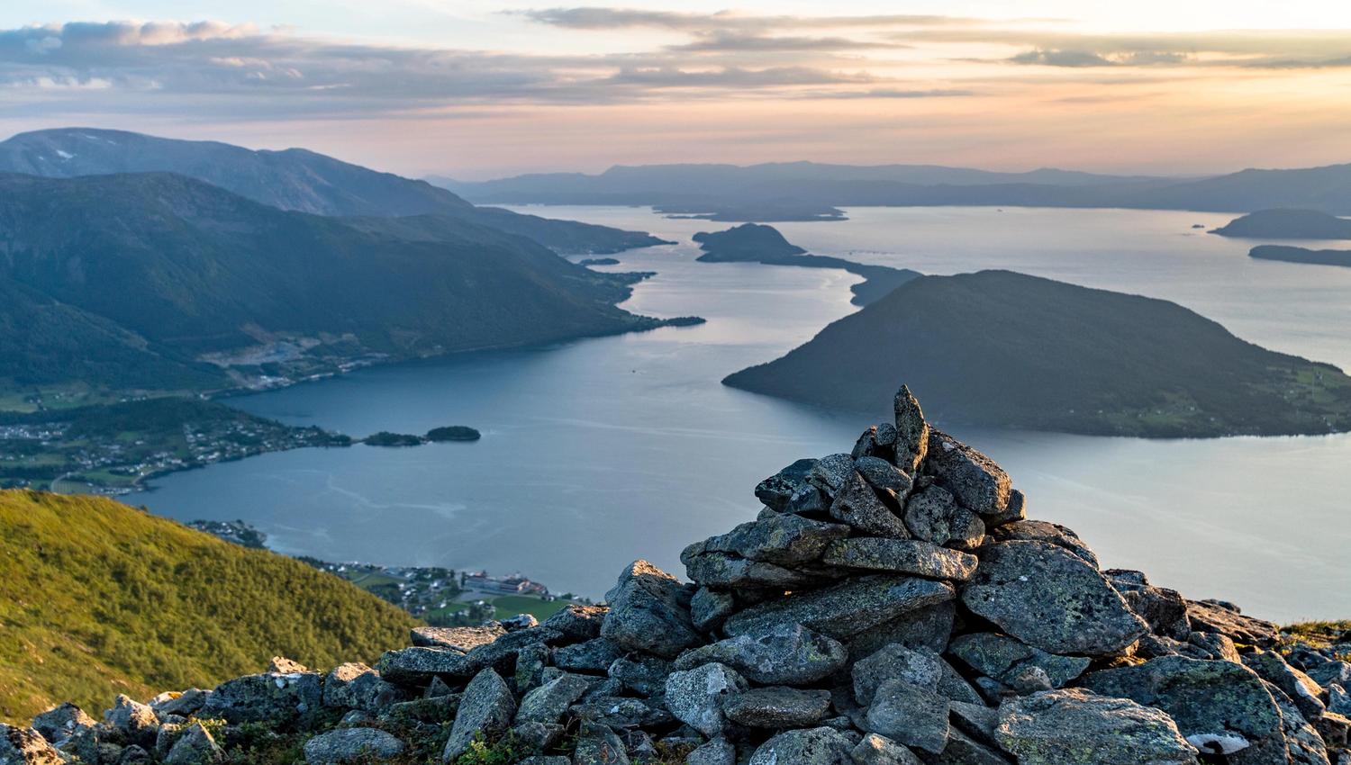 Panoramautsikt frå Malmangernuten over den djupe Hardangerfjorden og grøne øyar, med ein turgåar som nyt utsikta frå fjellkanten.