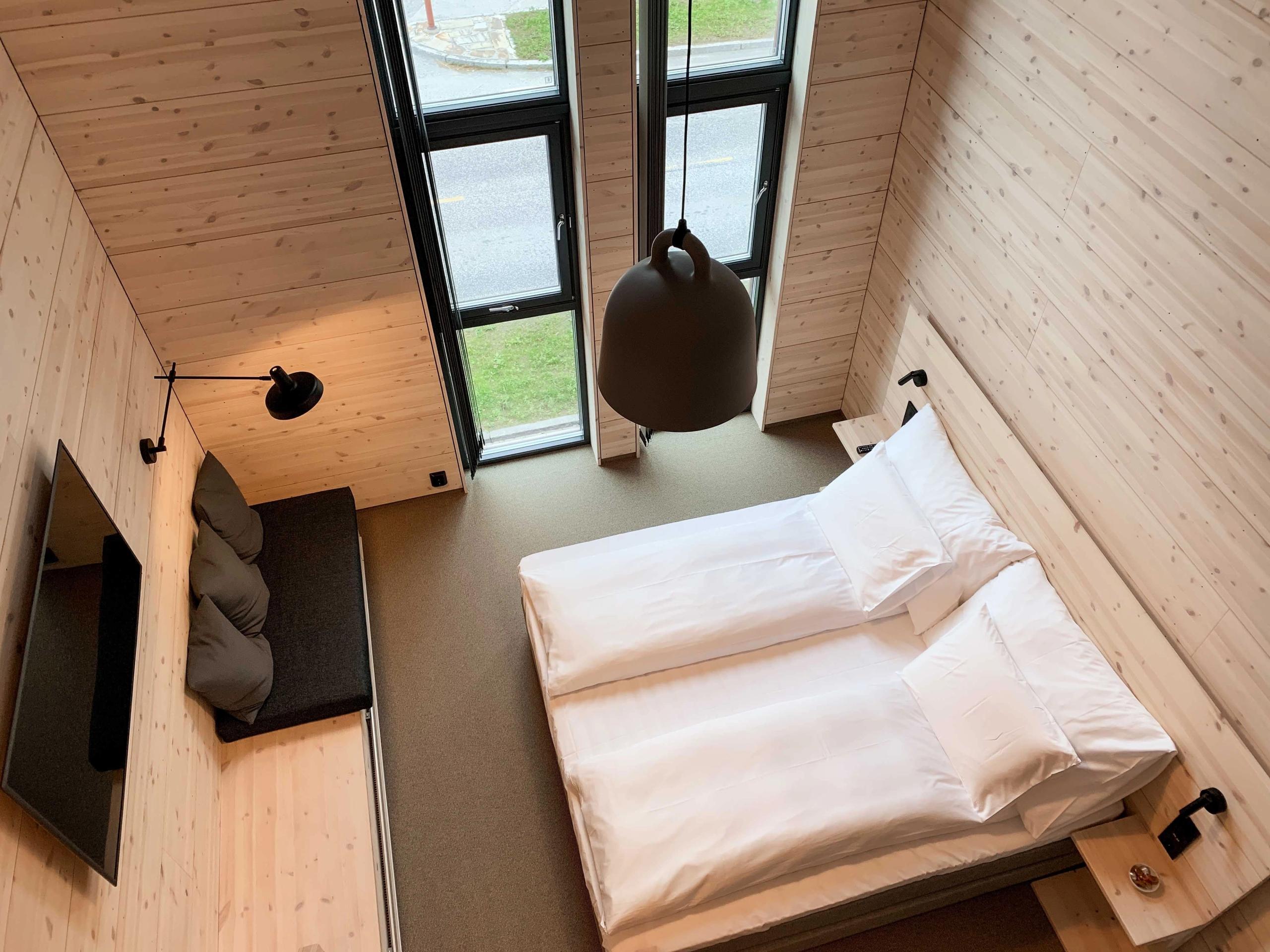Overhead view of a modern wood-paneled bedroom with twin beds, a hanging lamp, bench seating, and tall windows..