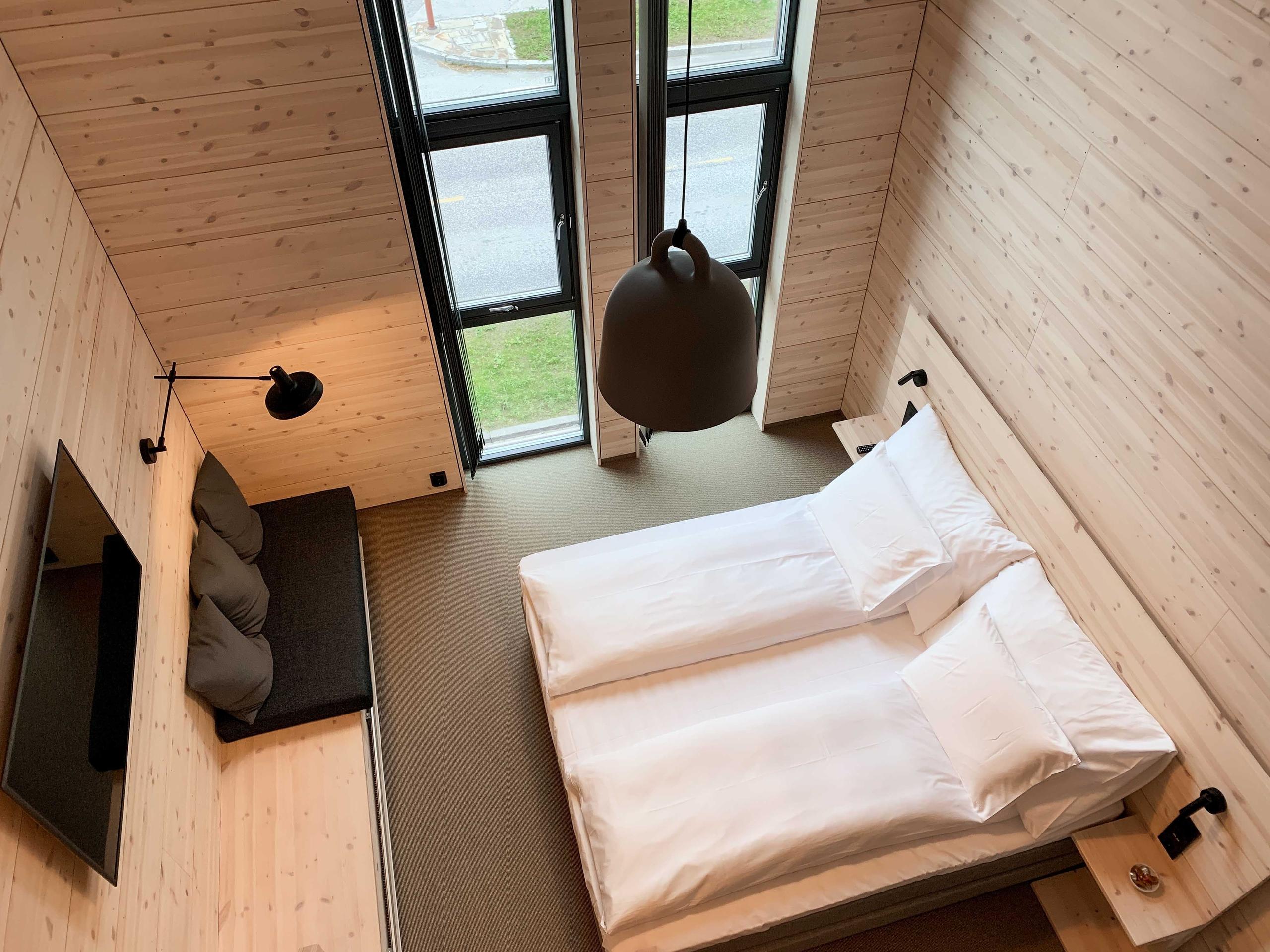 Overhead view of a modern wood-paneled bedroom with twin beds, a hanging lamp, bench seating, and tall windows..