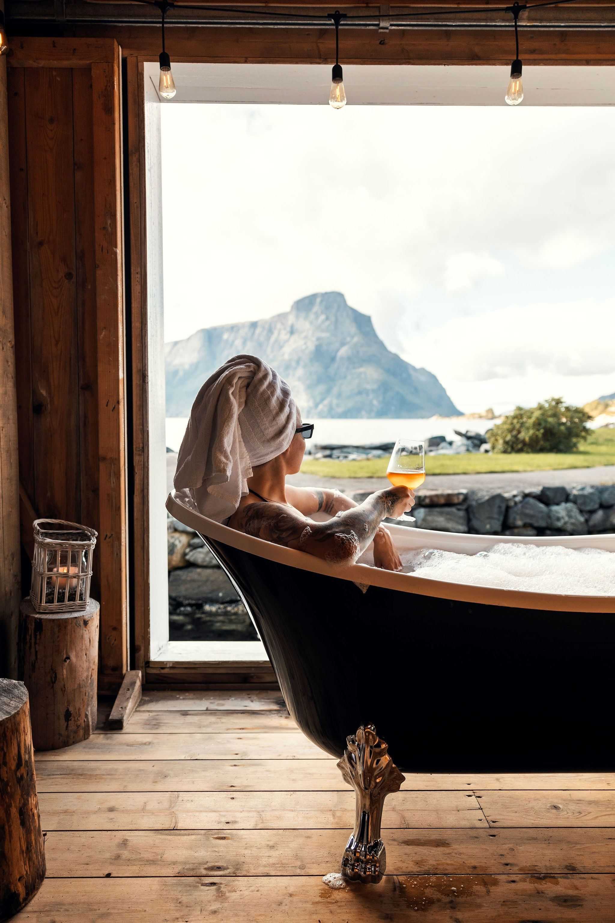 Person in a clawfoot tub with towel wrapped on head holds a drink, facing a mountain view through a large window at the Seahouse at Hornelen Panorama in Bremanger...
