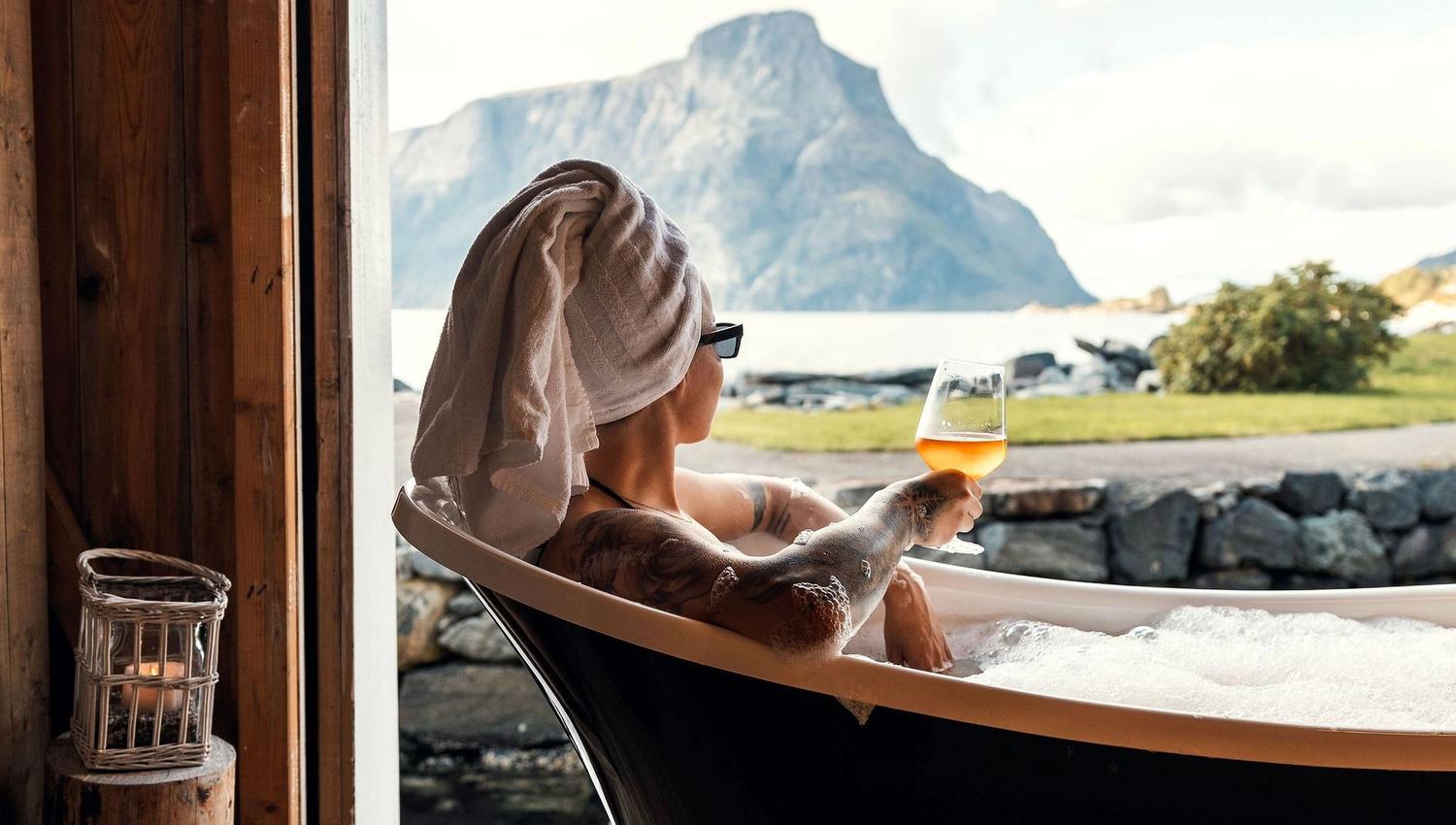 Person in a clawfoot tub with towel wrapped on head holds a drink, facing a mountain view through a large window at the Seahouse at Hornelen Panorama in Bremanger...