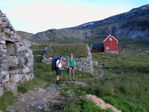 Hikers by a traditional red mountain cabin on Hardangervidda, surrounded by open alpine plateau scenery.