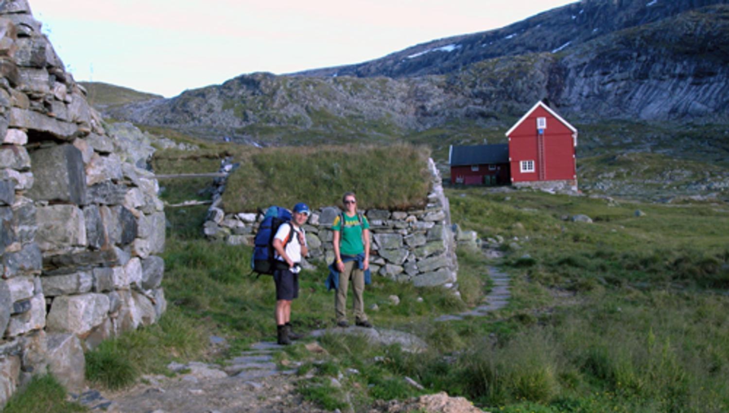 Hikers by a traditional red mountain cabin on Hardangervidda, surrounded by open alpine plateau scenery.