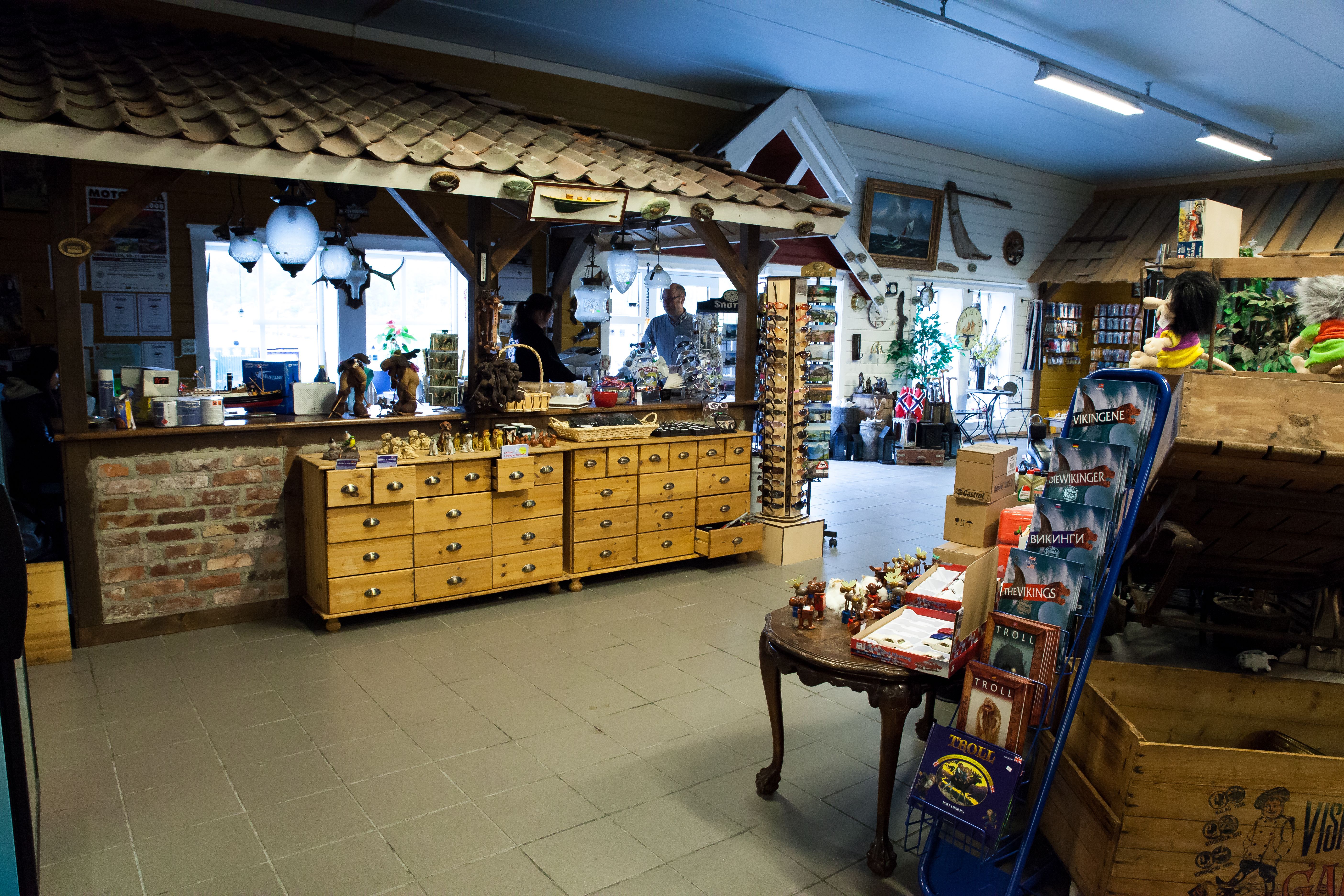 Checkout counter with a staff member; a man is about to pay. Lots of items all around: trolls, books about Vikings, sunglasses, moose figurines, and s