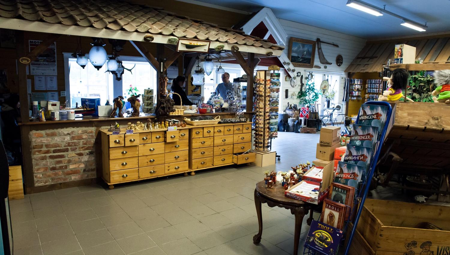 Checkout counter with a staff member; a man is about to pay. Lots of items all around: trolls, books about Vikings, sunglasses, moose figurines, and s
