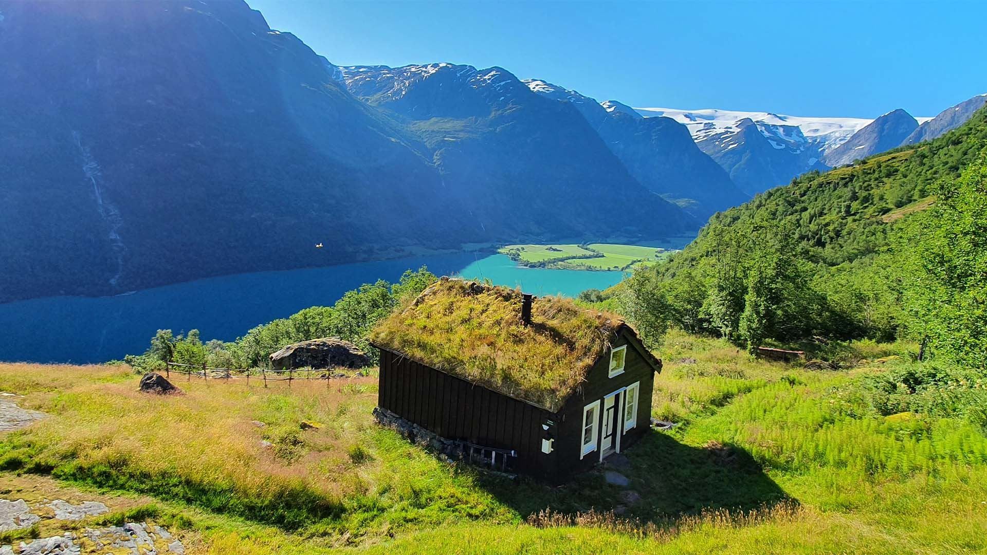 Small dark cabin with a grass roof on a sunny hillside, overlooking a blue lake and snow-capped mountains.