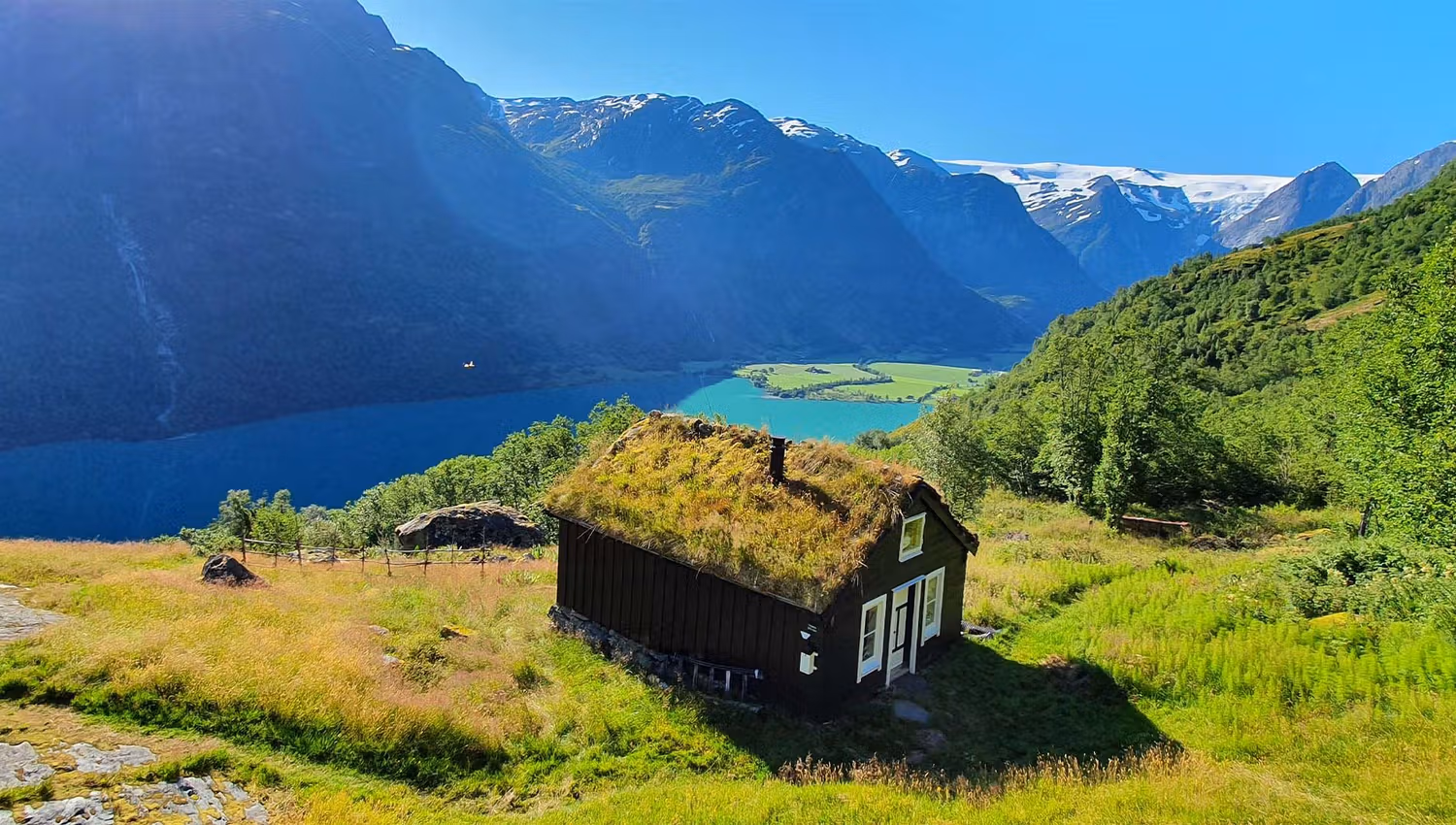 Small dark cabin with a grass roof on a sunny hillside, overlooking a blue lake and snow-capped mountains.