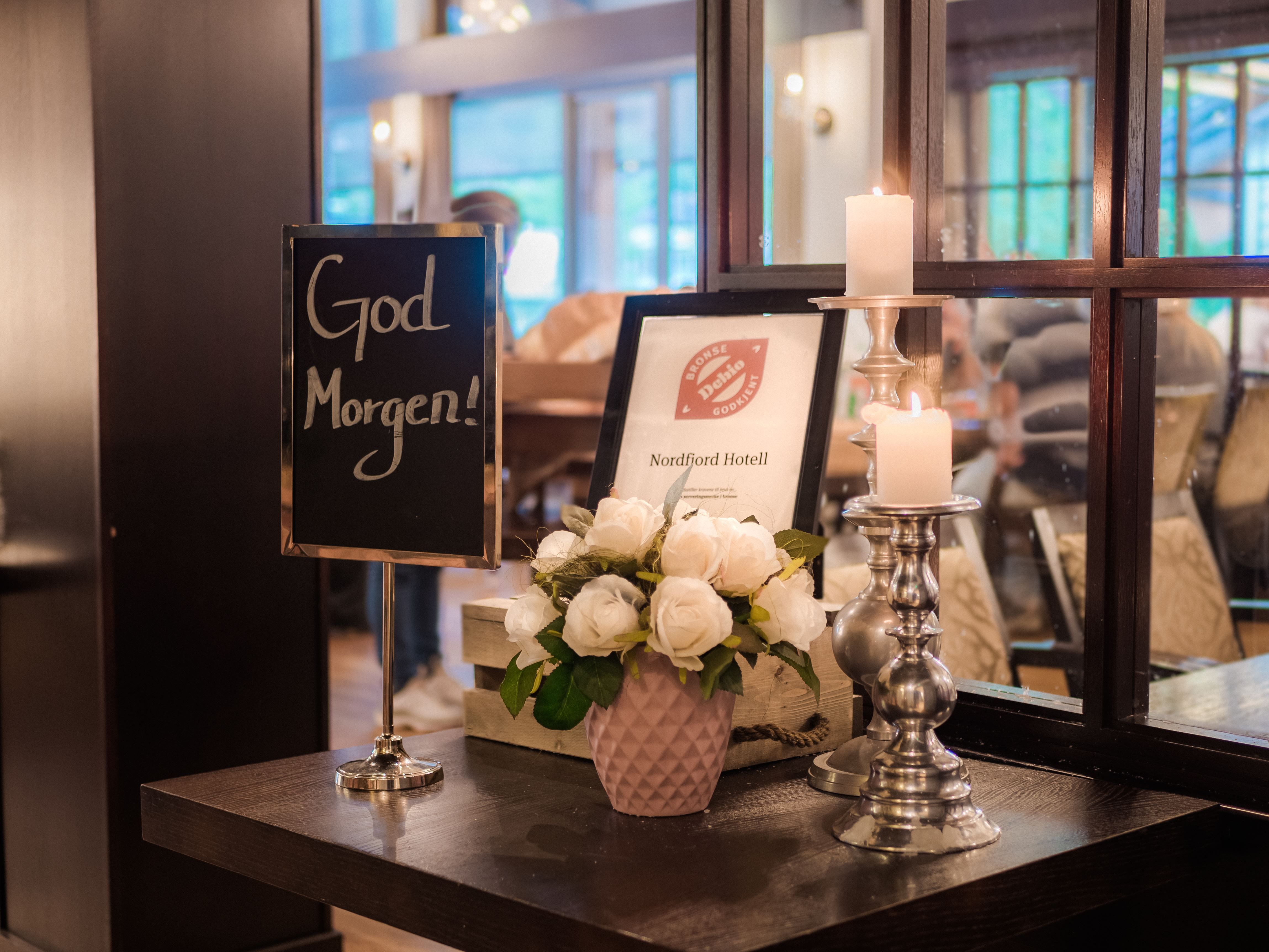 Table display with “God Morgen!” sign, framed hotel certificate, white roses in a vase, and two lit candles by a window.