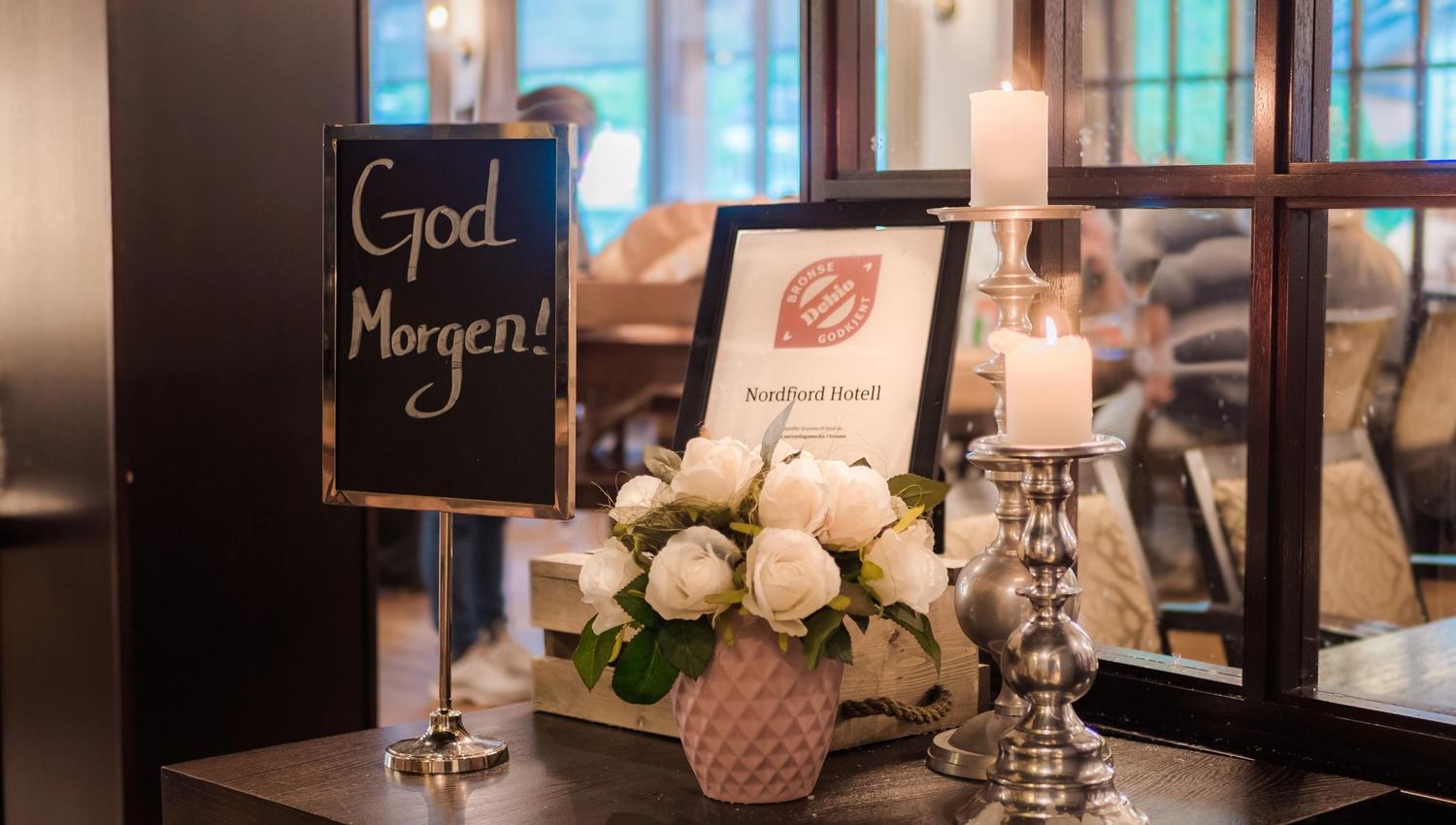 Table display with “God Morgen!” sign, framed hotel certificate, white roses in a vase, and two lit candles by a window.