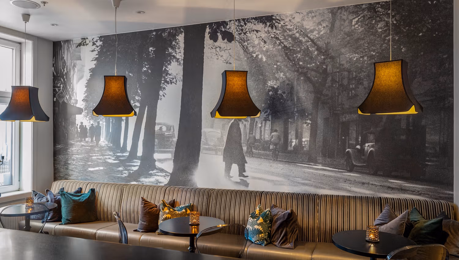 langsofa med puter. Fototapet med bymotiv. Brune lampetter over bordet. Saga Hotel Oslo. Foto