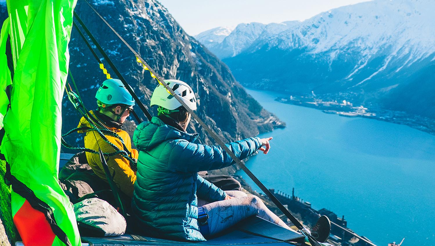 To personar sit på ein portaledge ved fjorden i Hardanger, med majestetiske fjell i bakgrunnen. Ein unik overnattingoppleving i fjellveggen.