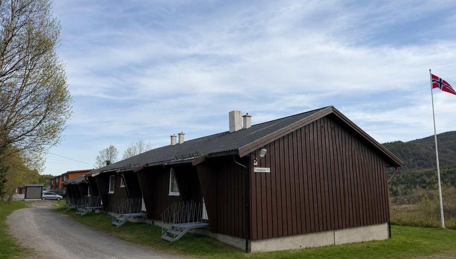 Long brown wooden building with small porches beside a gravel road, under a cloudy sky with a flagpole at right.