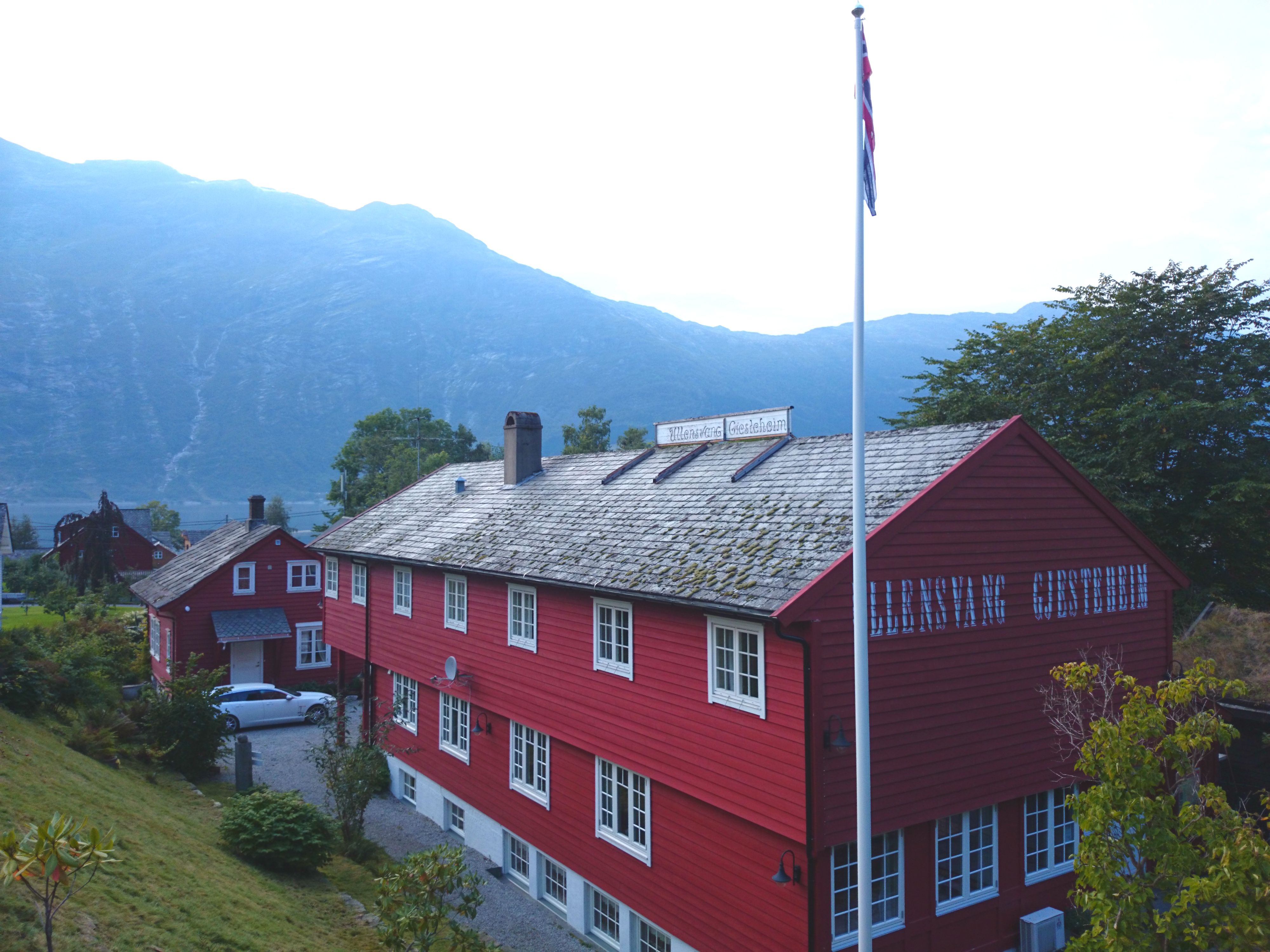 Ullensvang Gjesteheim viewed from the courtyard, featuring its red wooden building, Norwegian flag and mountain backdrop.